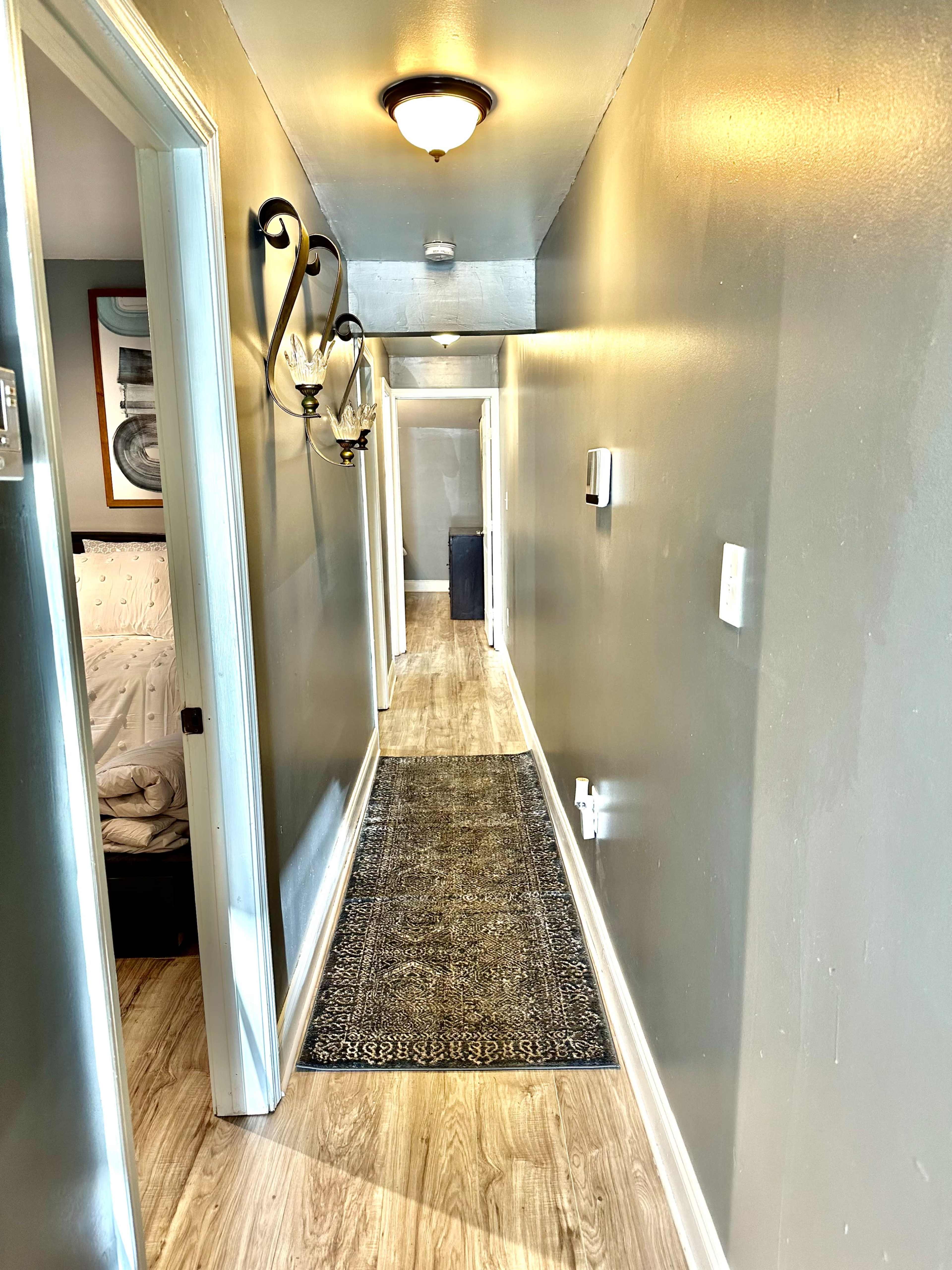 The image shows a narrow hallway with a decorative rug, light fixtures on the walls, and doors leading to other rooms.