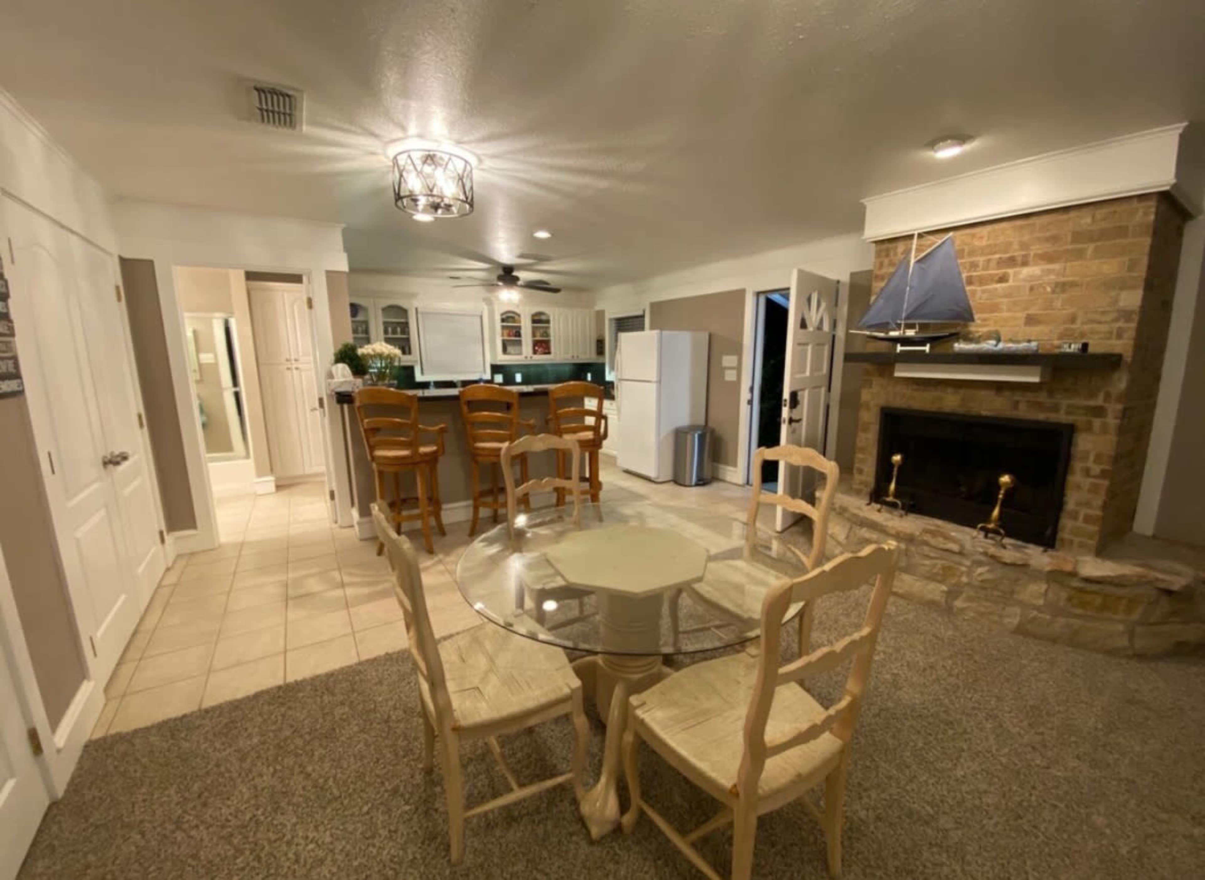 A light-colored kitchen and dining area with a round glass table, four chairs, a stone fireplace, and a bar with high stools.