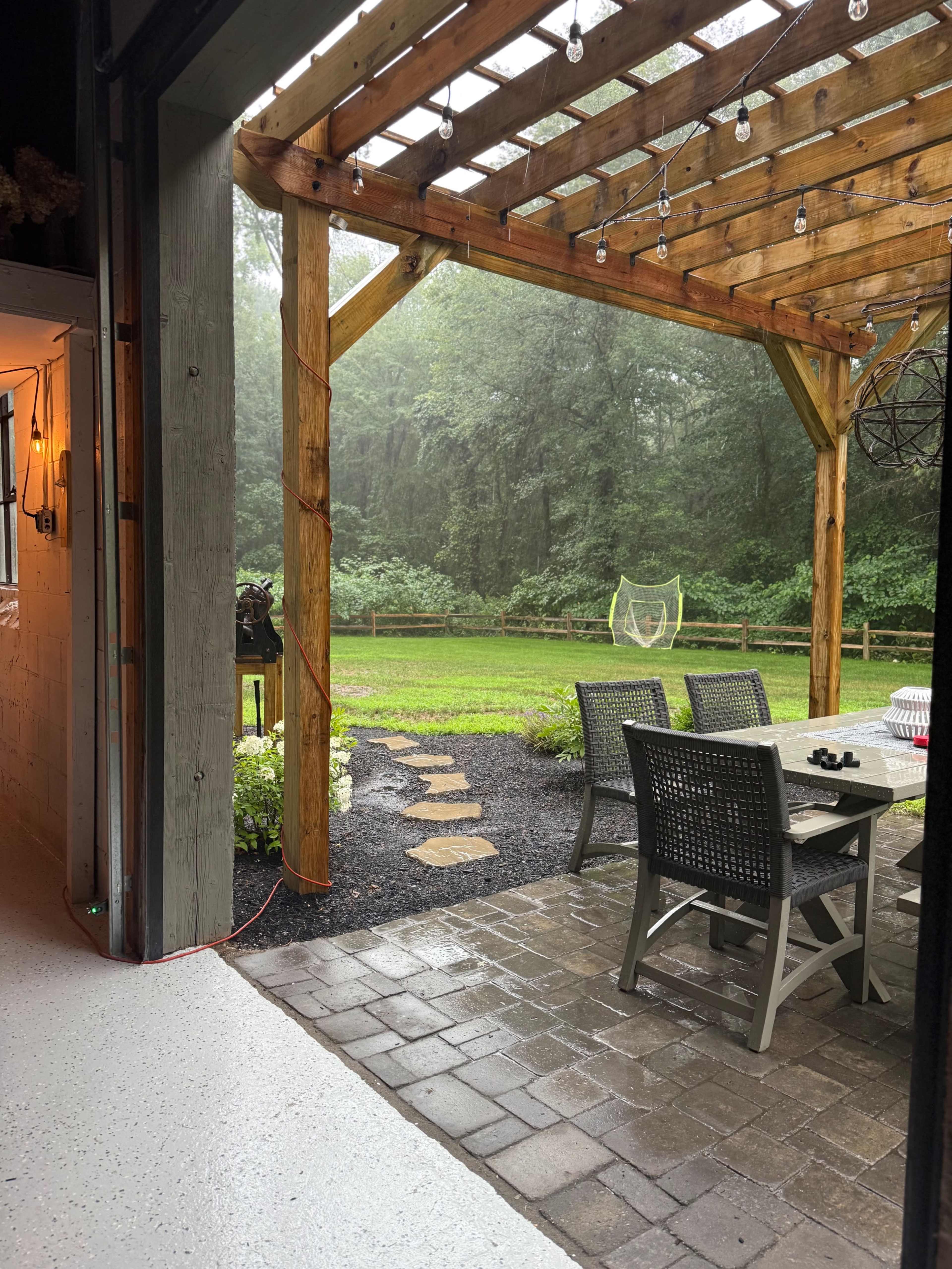 A patio with wooden beams and hanging lights overlooks a grassy yard surrounded by trees on a rainy day.