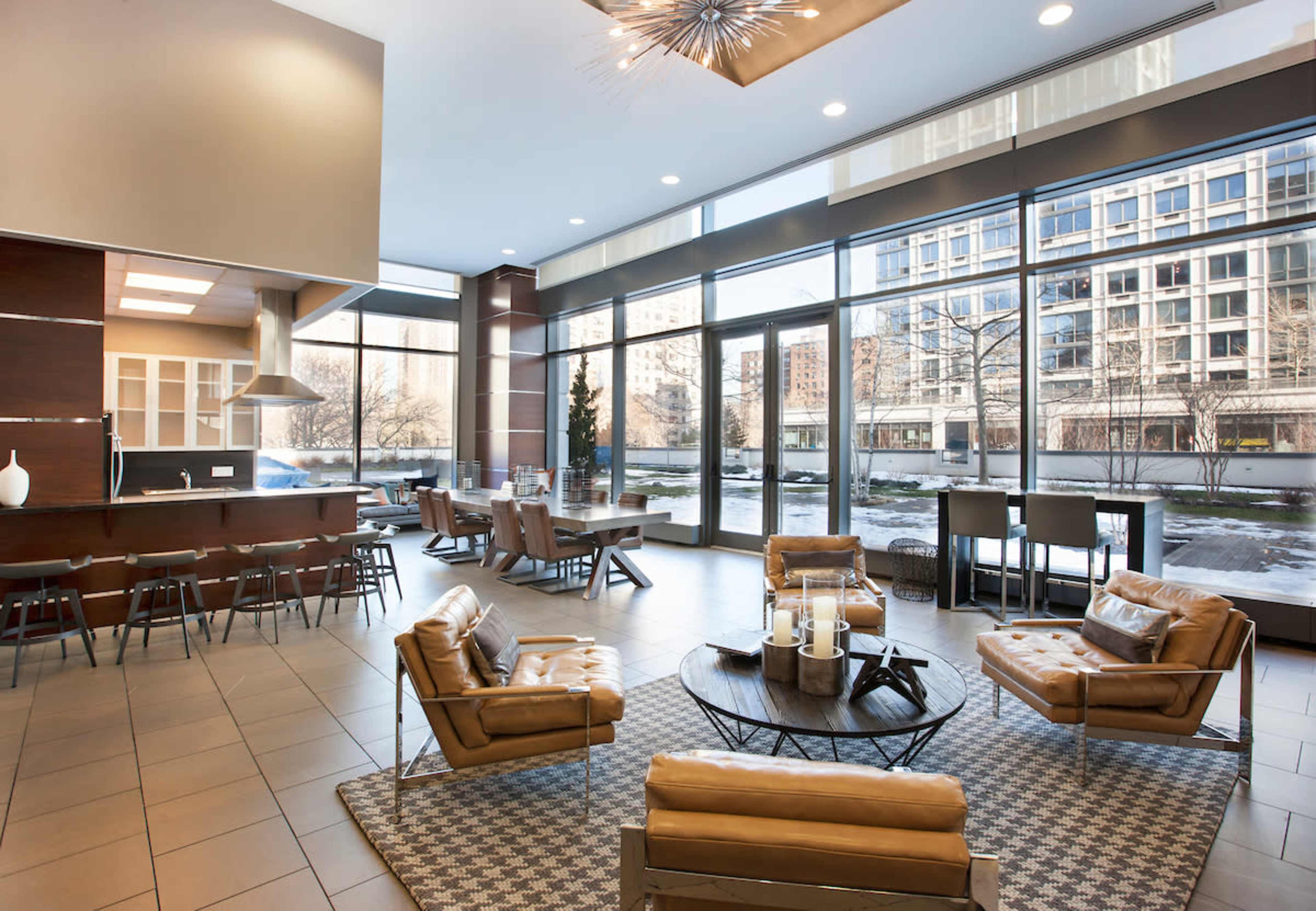 The image shows a modern lobby with large windows, featuring leather seating, a round coffee table, and a kitchen area in the background.