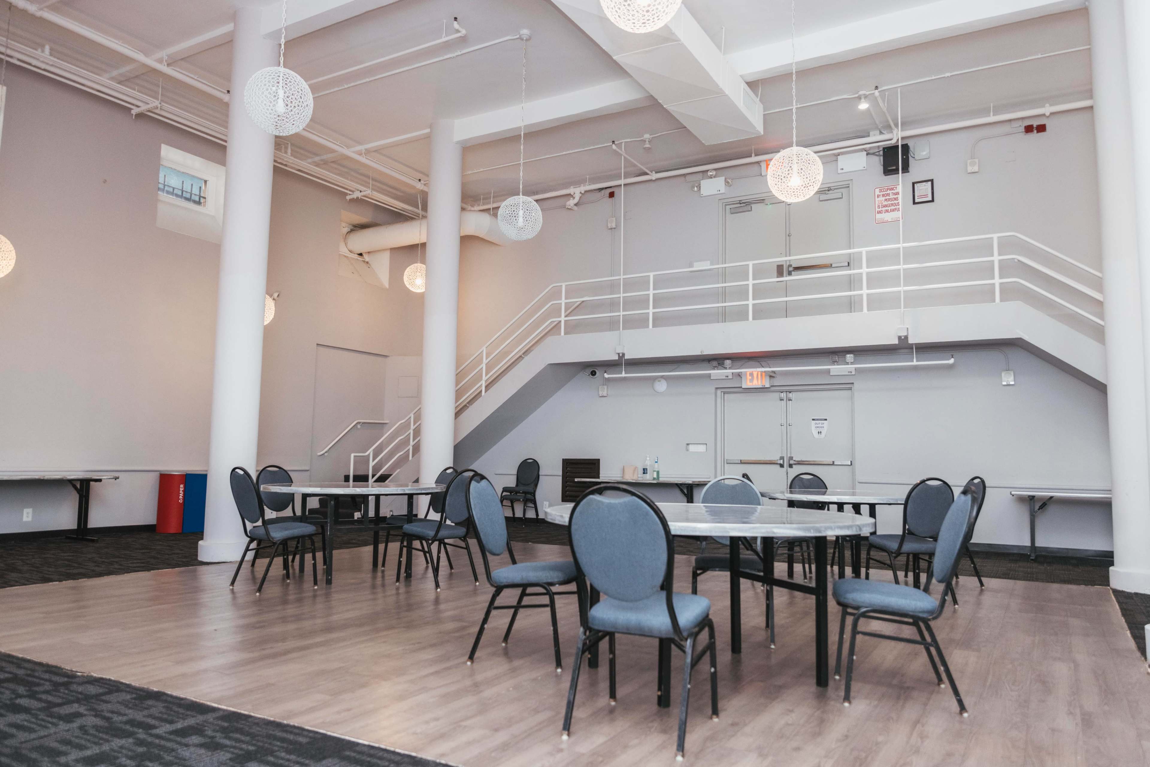 The image shows a spacious, modern room with several round tables and chairs arranged on a hardwood floor, complemented by a staircase in the background.