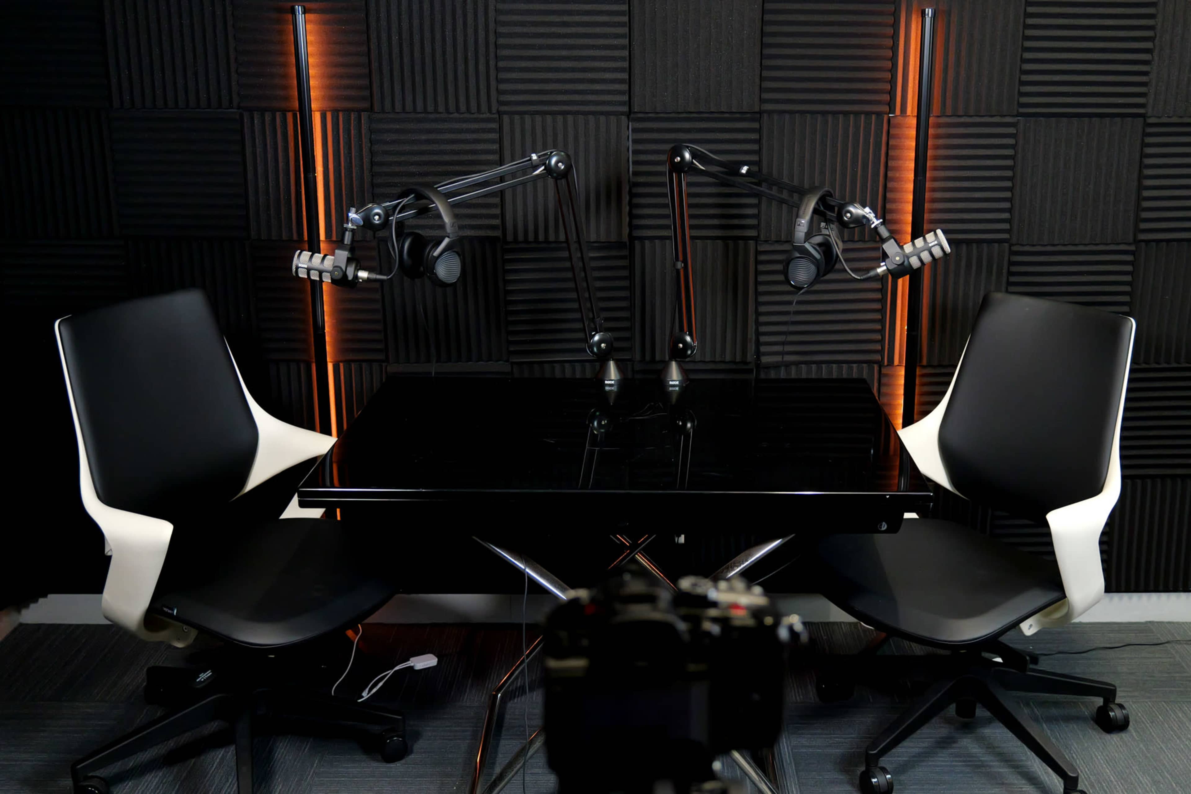 A podcast studio setup features a black table with two modern chairs and two microphone arms, set against a textured wall with orange lighting.