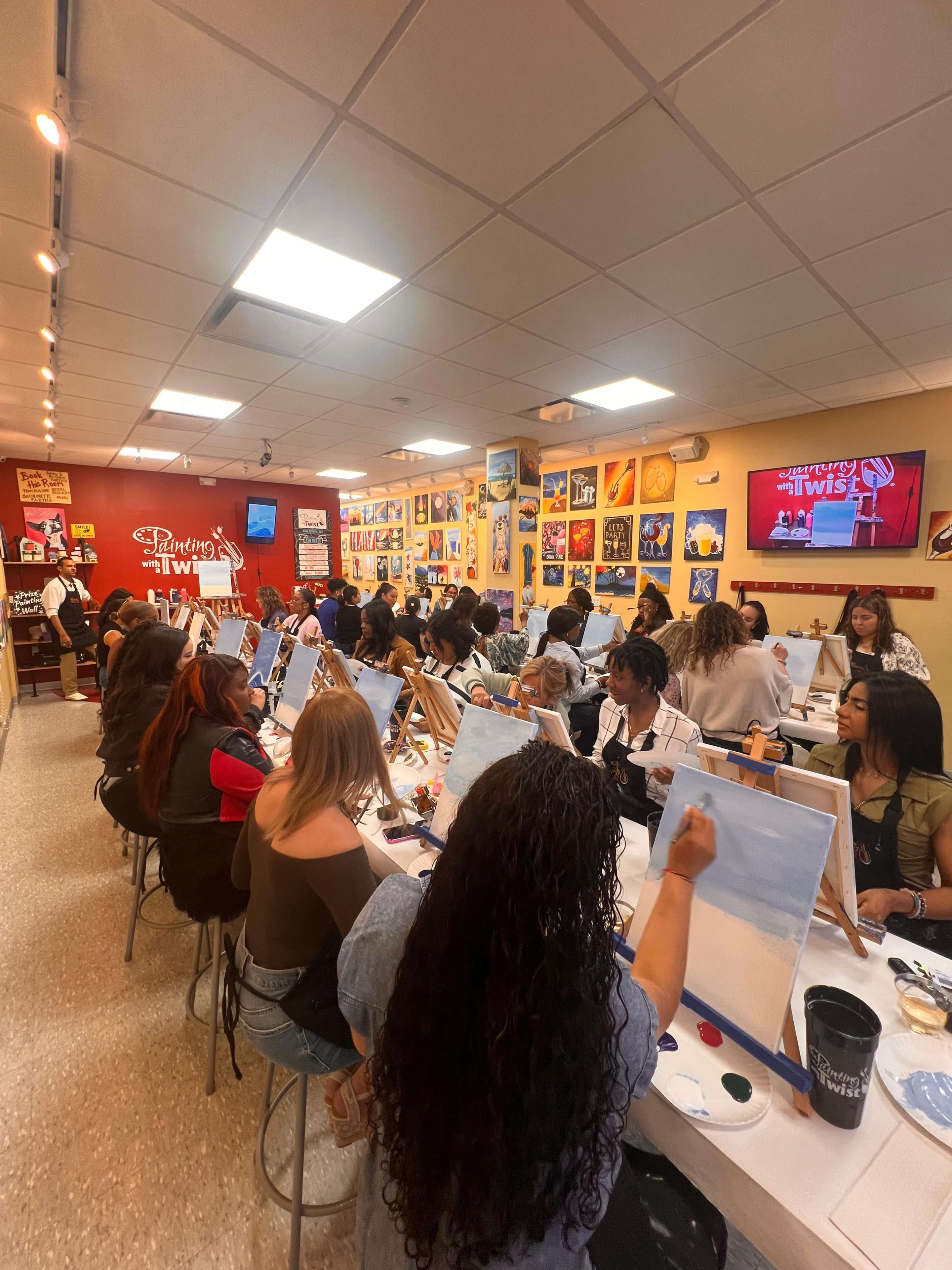 A group of people sits at easels in an art studio, each painting on canvases during a guided painting event.