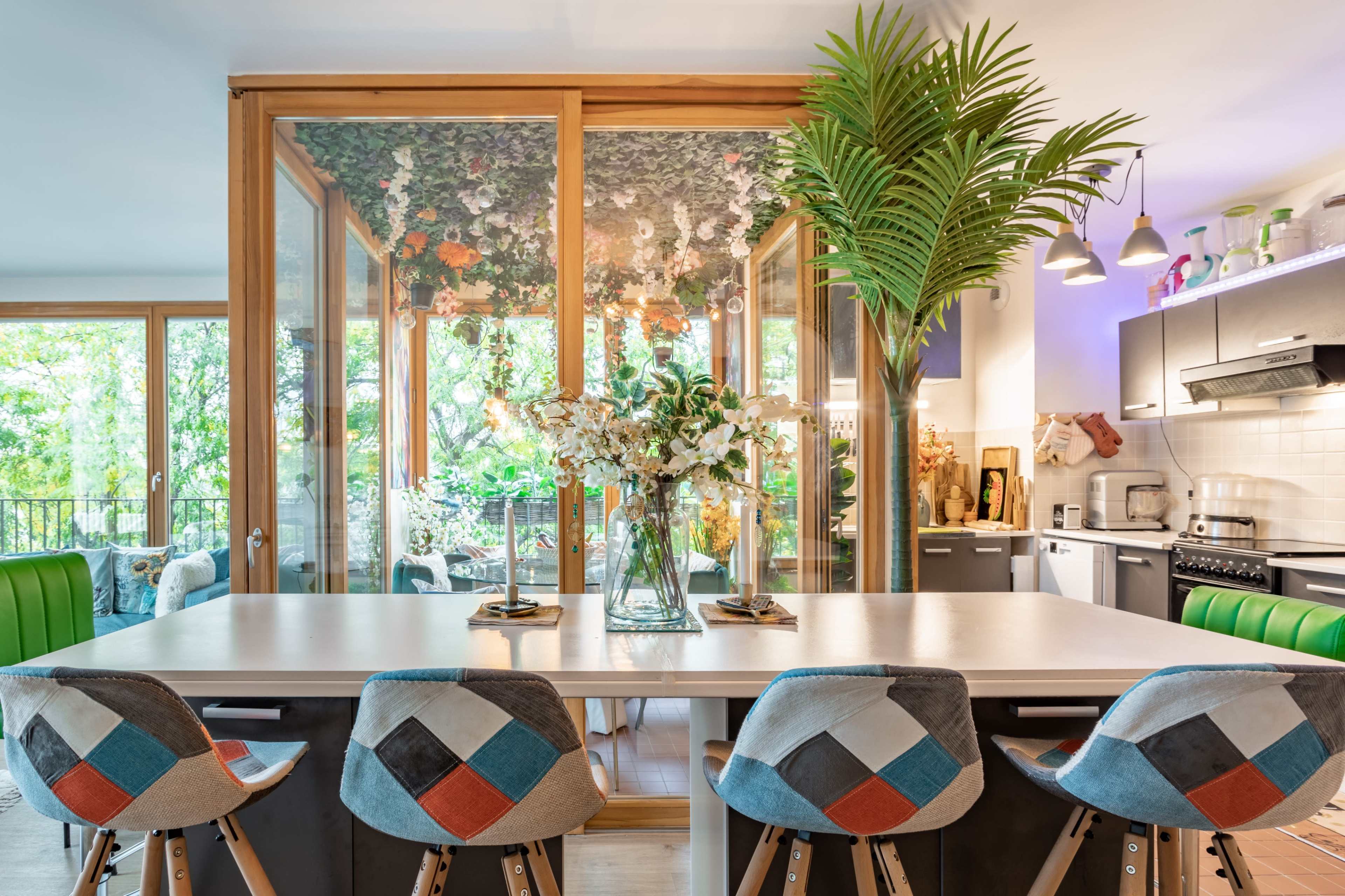 A modern kitchen with a dining area featuring a large white table, colorful upholstered bar stools, and a glass-enclosed room filled with plants and flowers.