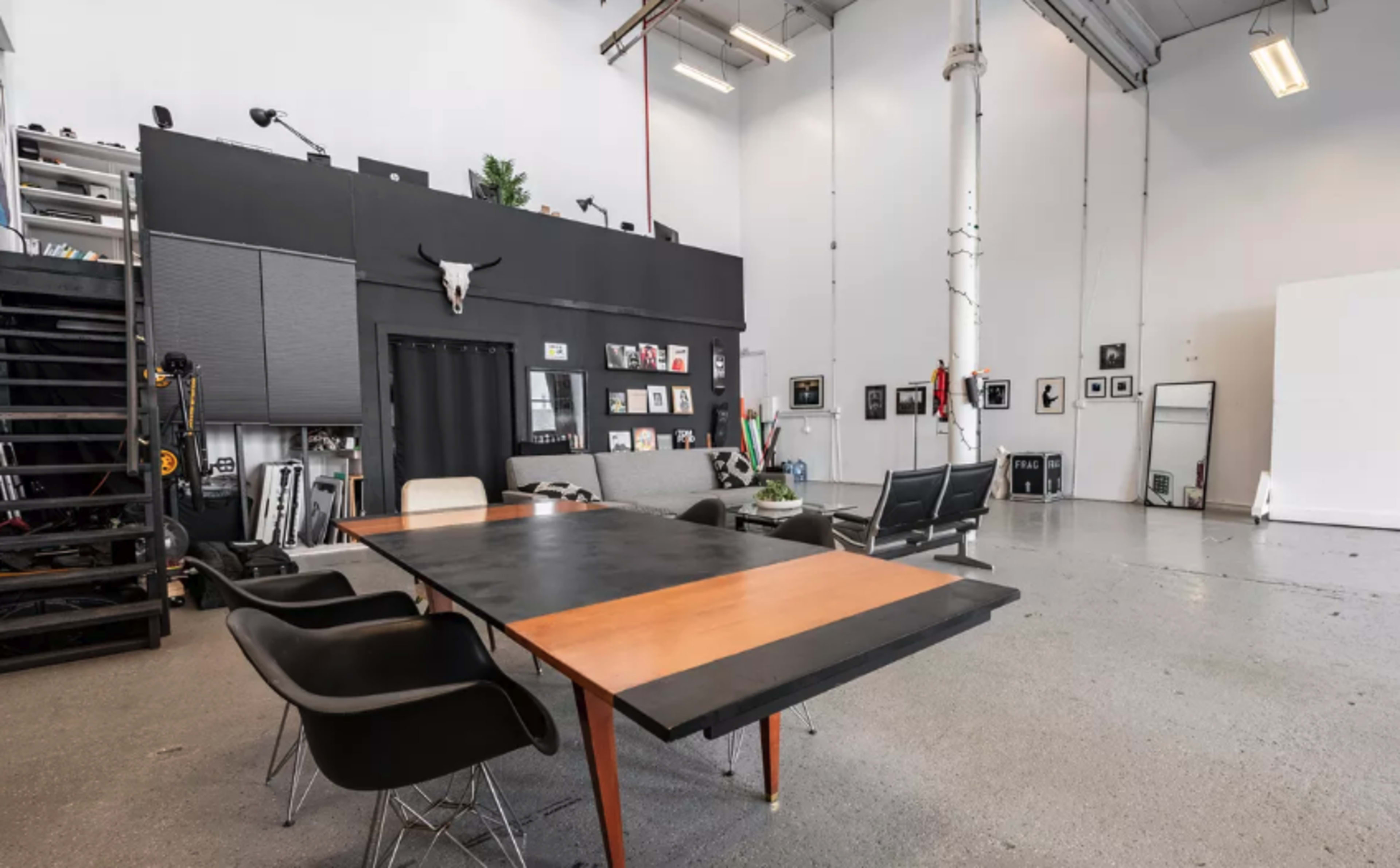 A spacious studio with a large table and chairs, black walls adorned with framed pictures, and a staging area featuring a blank backdrop.