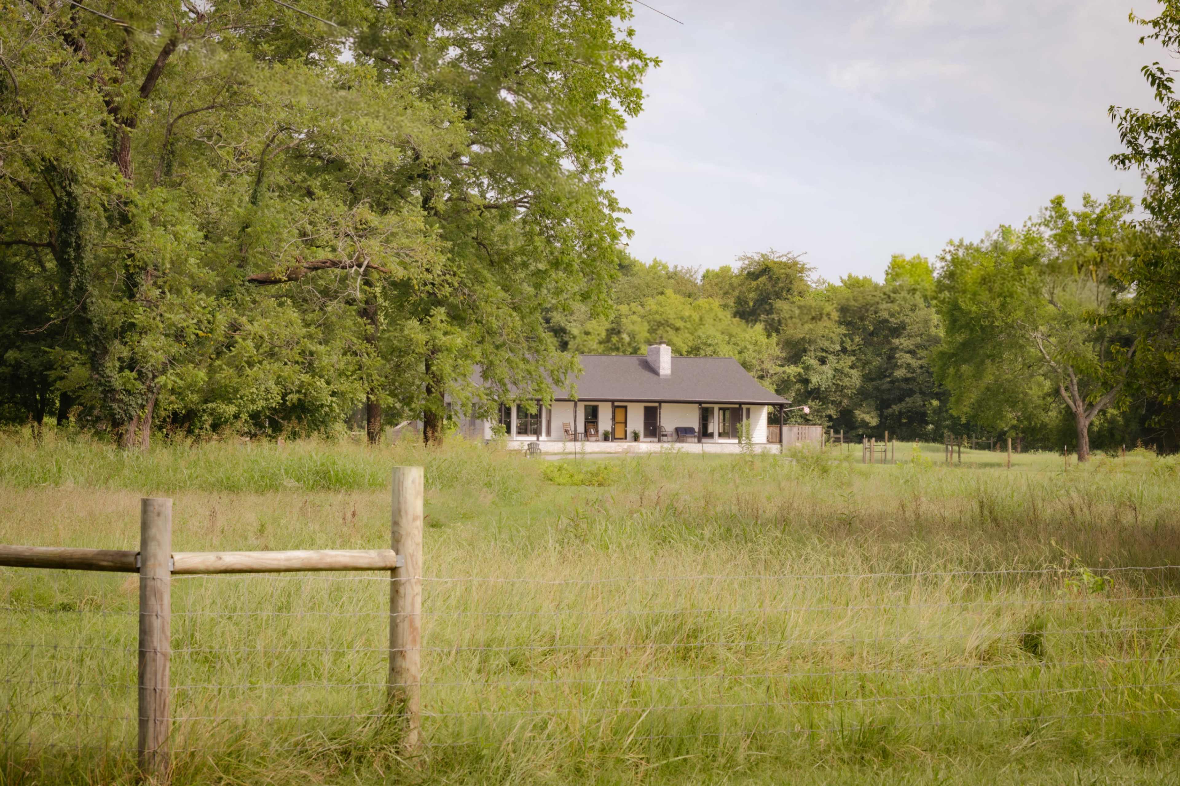 A single-story house with a porch is situated among tall grass and trees, with a wooden fence in the foreground.
