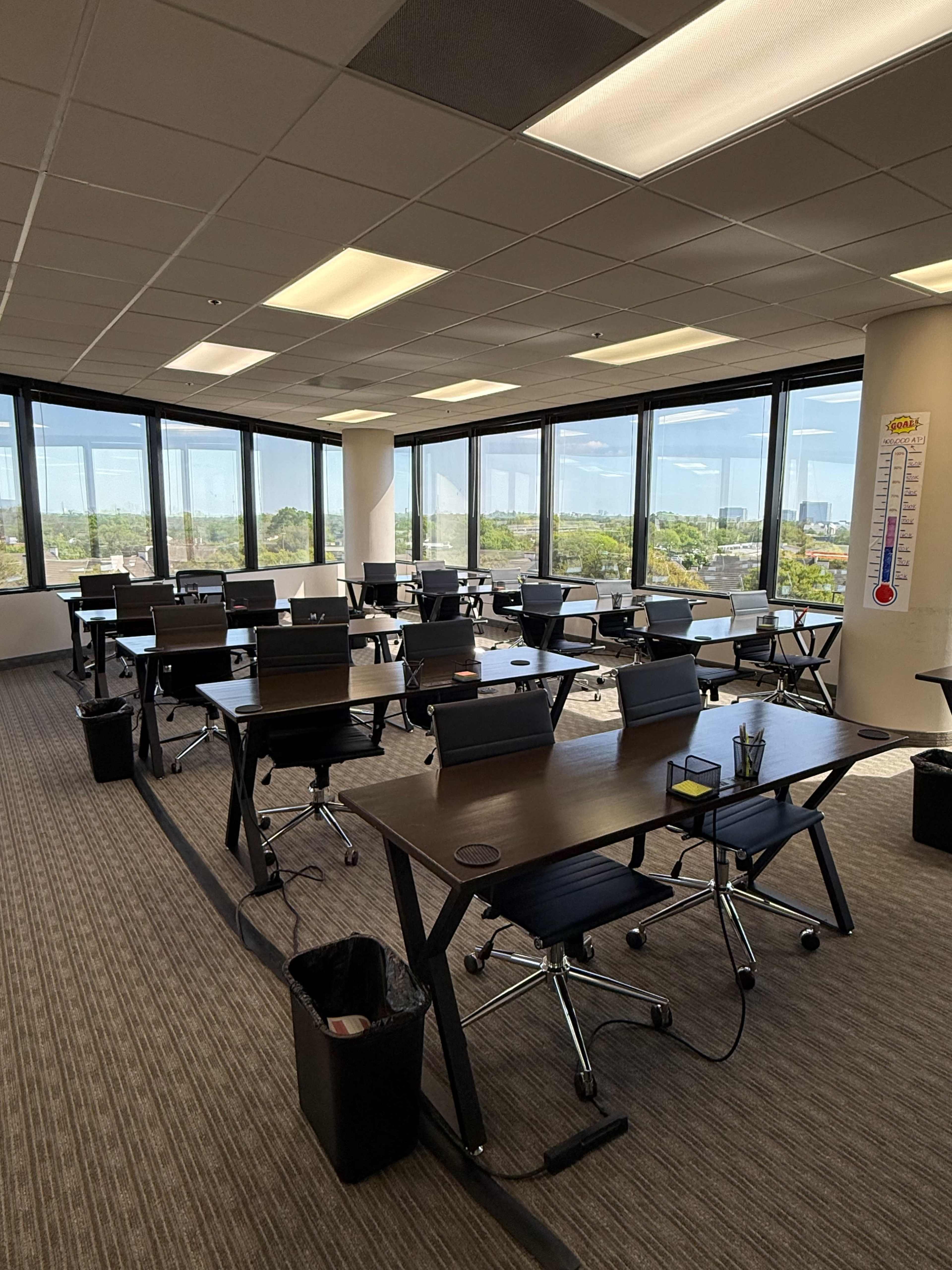 The image shows a spacious office environment with multiple desks arranged in rows, large windows allowing natural light, and a view of greenery outside.