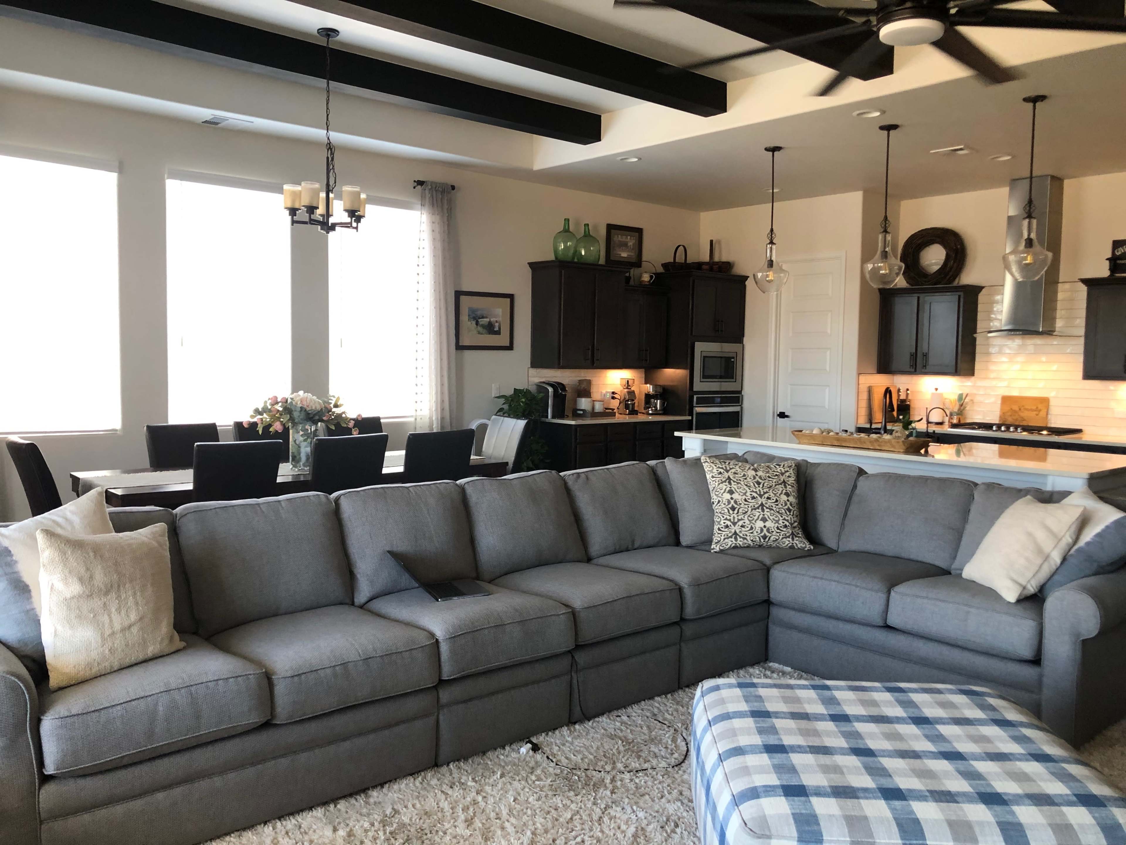 The image shows a spacious living room with a gray sectional sofa, light-colored walls, large windows, and an adjoining dining area.