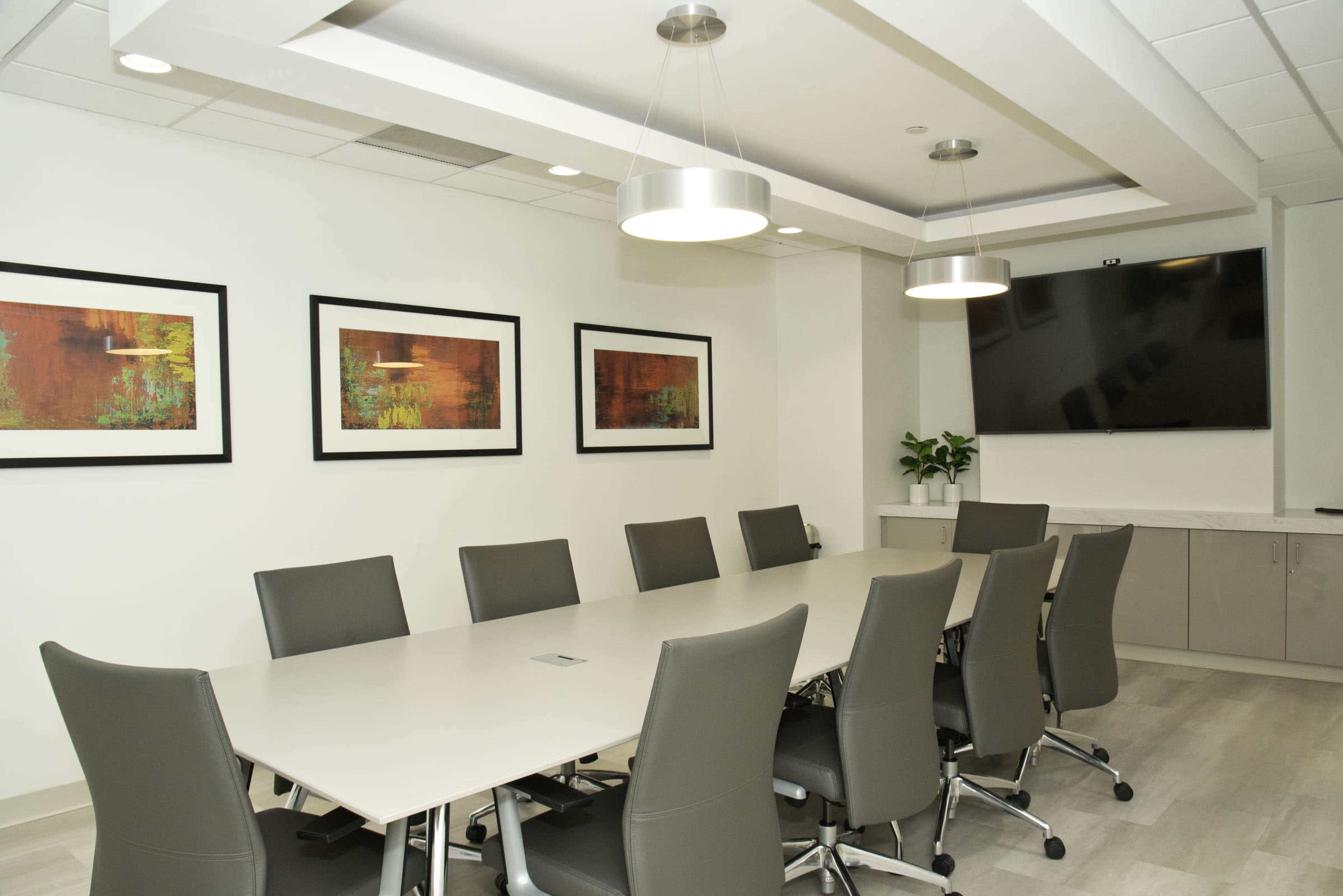 A conference room features a long table surrounded by gray chairs, with framed artwork on the walls and a television mounted in one corner.