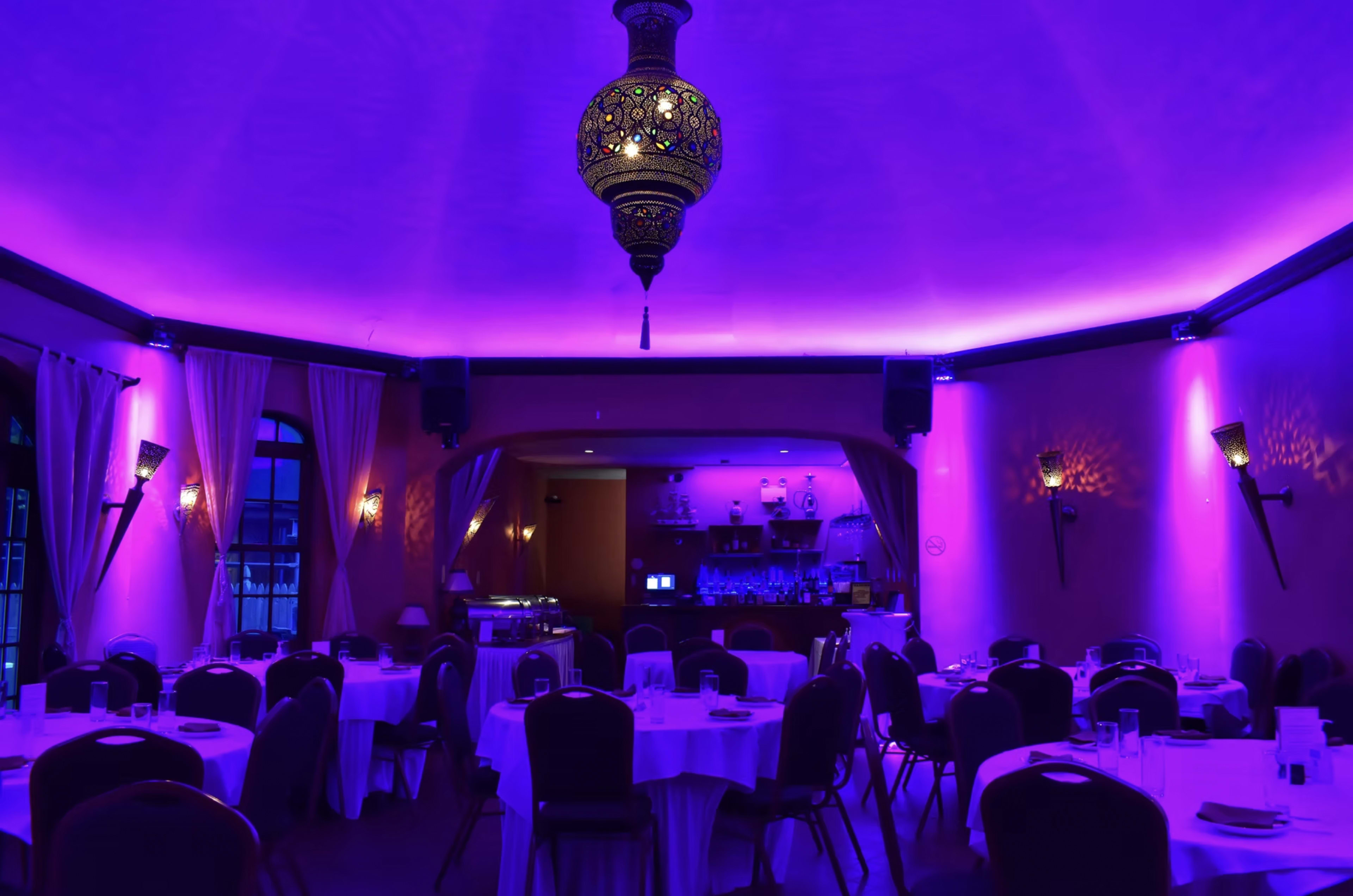 The image shows a dimly lit restaurant interior featuring round tables with white tablecloths, purple lighting, and an ornate lantern hanging from the ceiling.