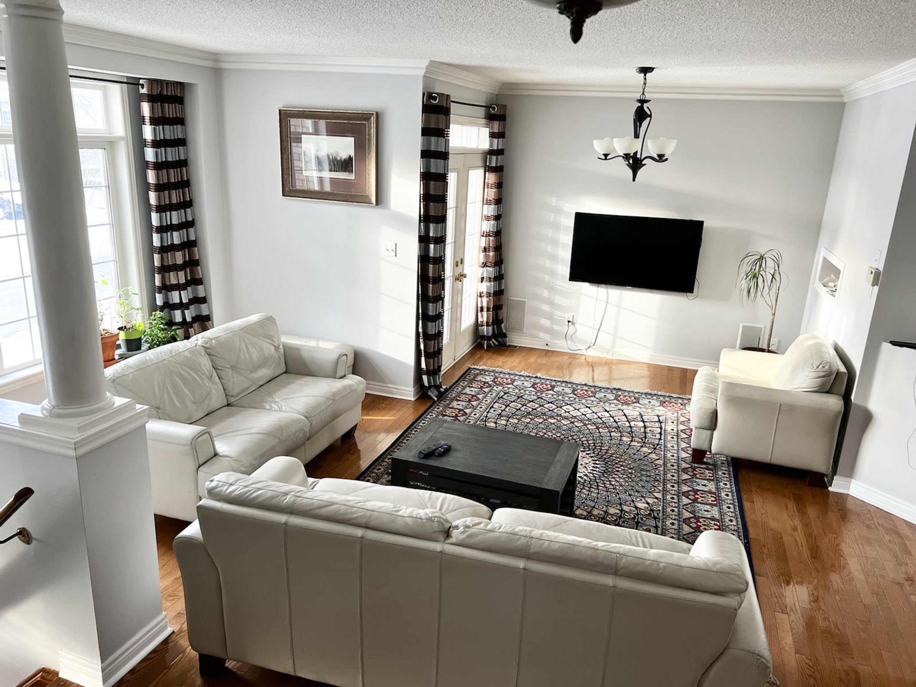 The living room features two white leather sofas, a black coffee table, and a television mounted on the wall, all arranged around a patterned area rug.