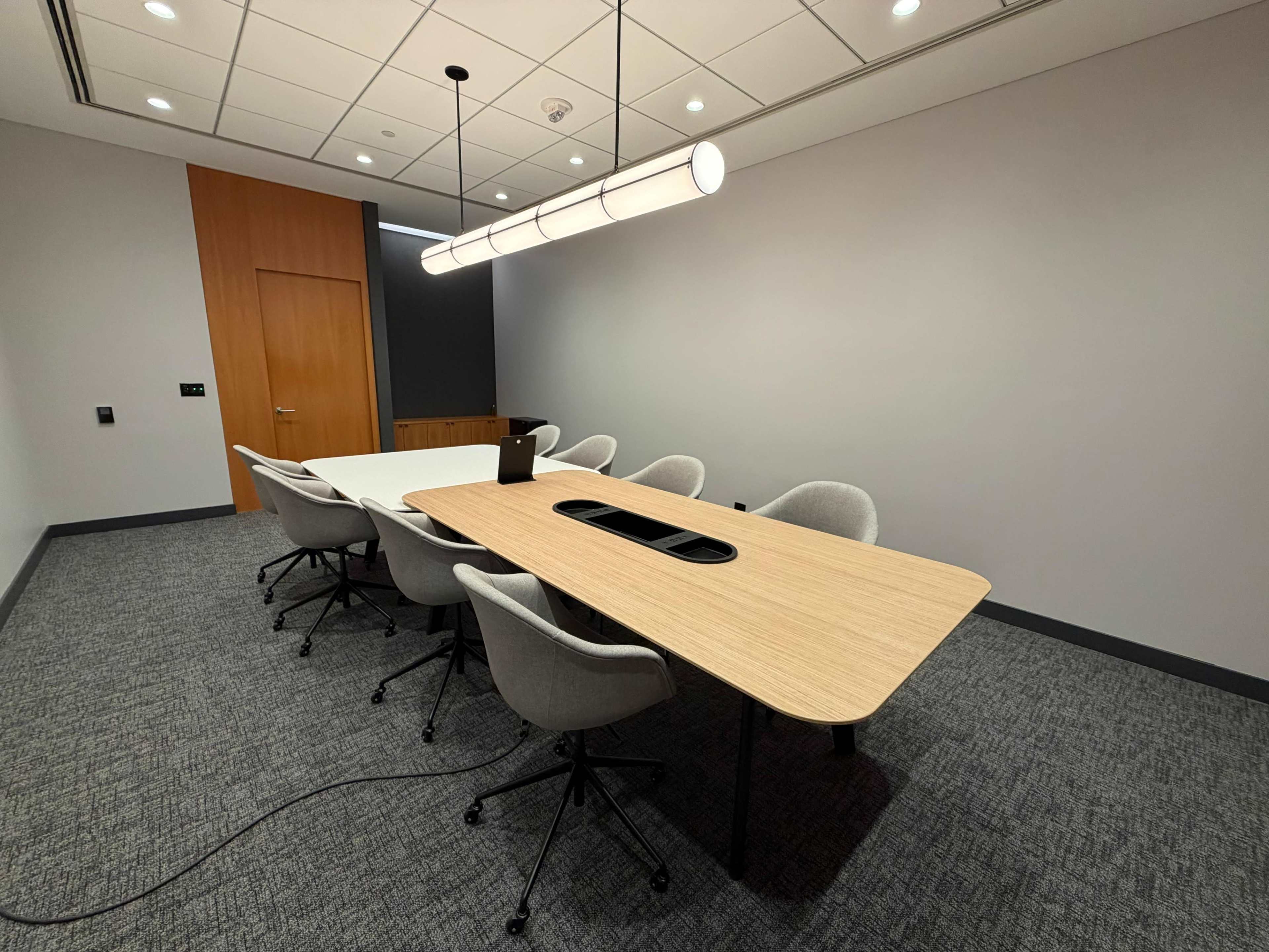 A modern conference room features a large wooden table surrounded by gray upholstered chairs and a central light fixture.
