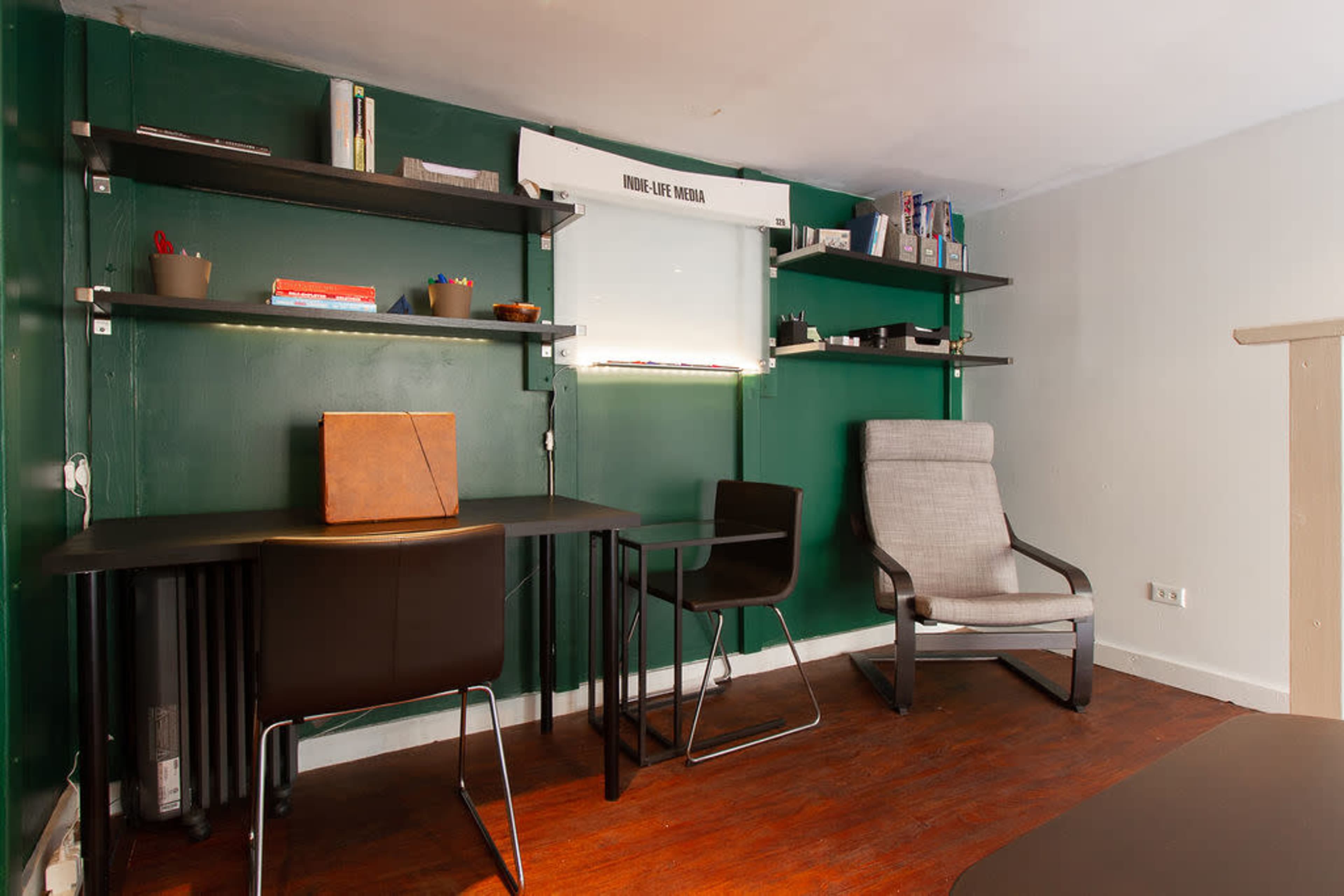 The image shows a small room with a green wall, a wooden floor, a desk with a chair, and shelves holding various items.