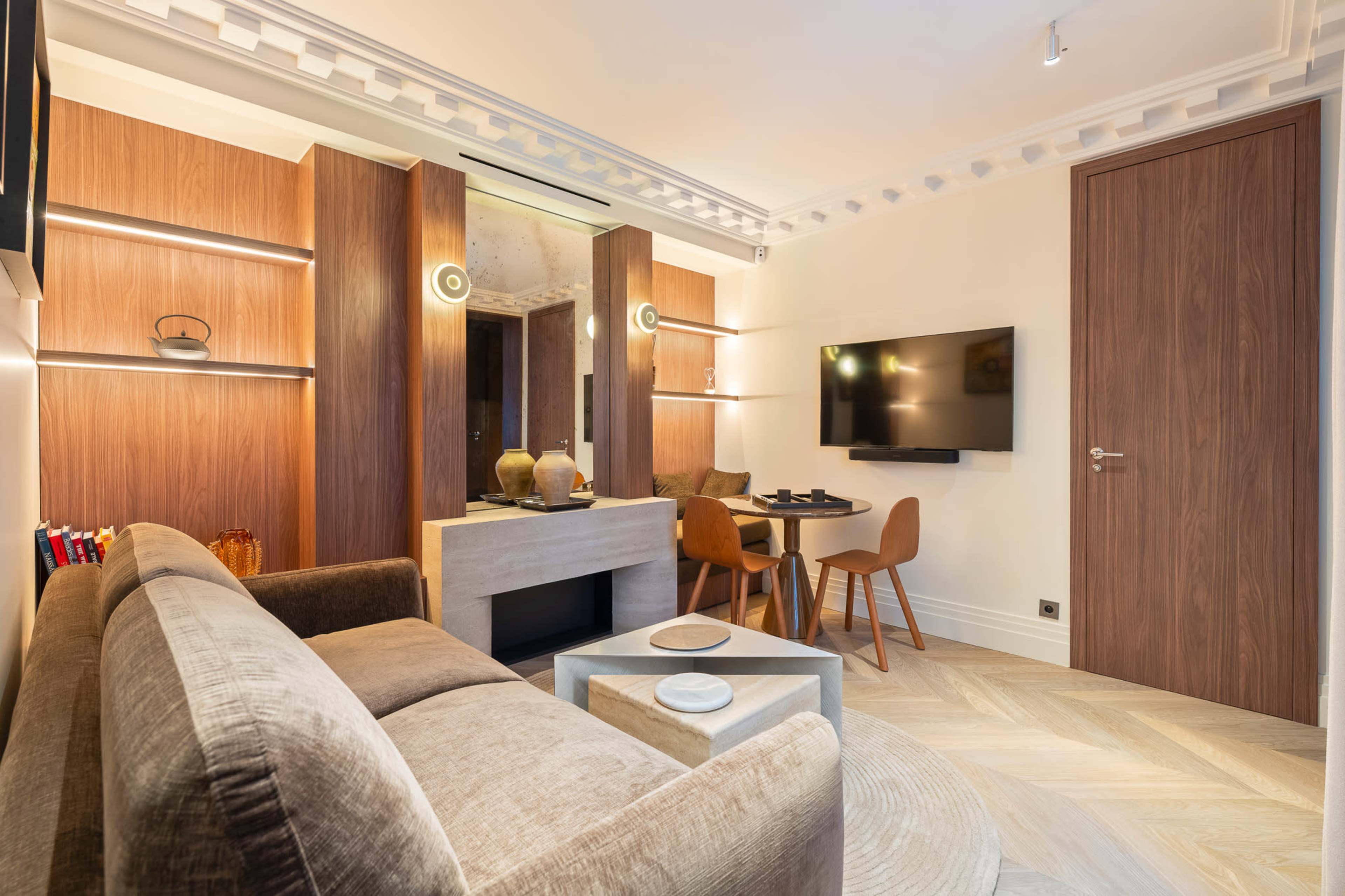 The image shows a stylish living area featuring a modern sofa, a wall-mounted television, and a small dining table with chairs.