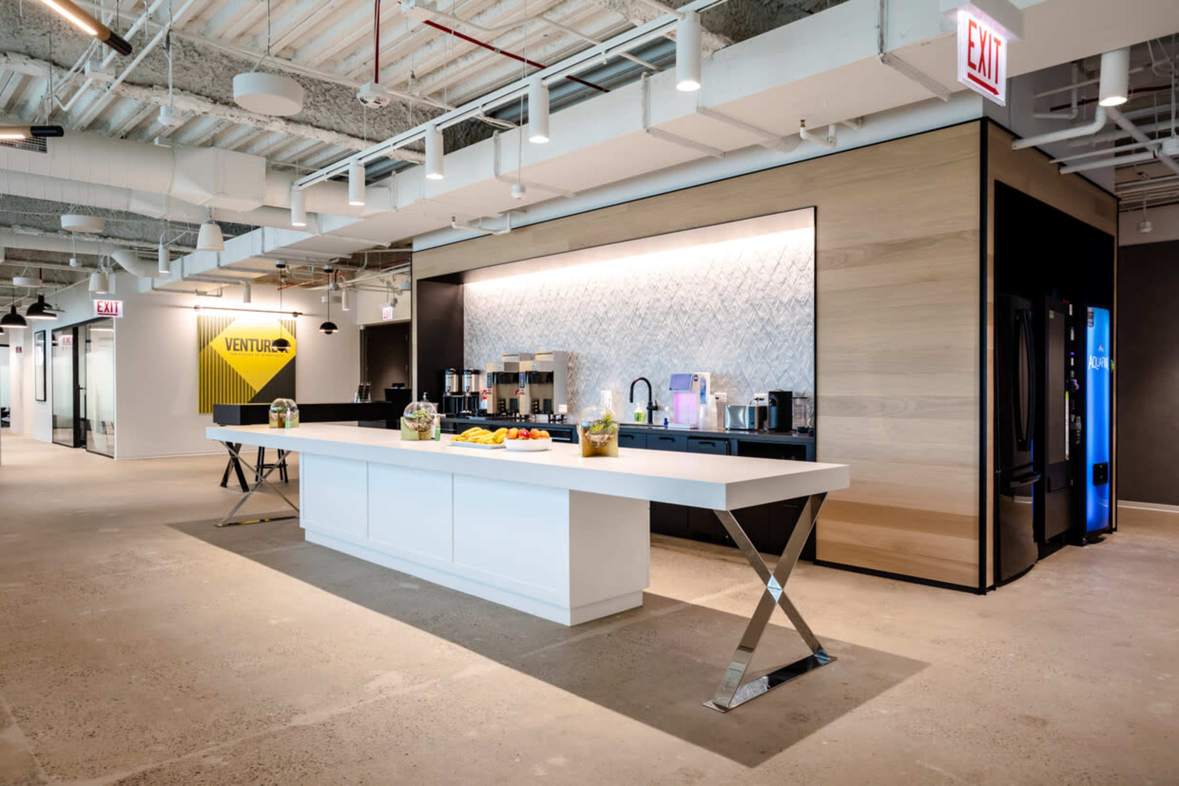 The image shows a modern office kitchen area featuring a long white countertop, coffee machines, and a variety of fruit displayed on top.