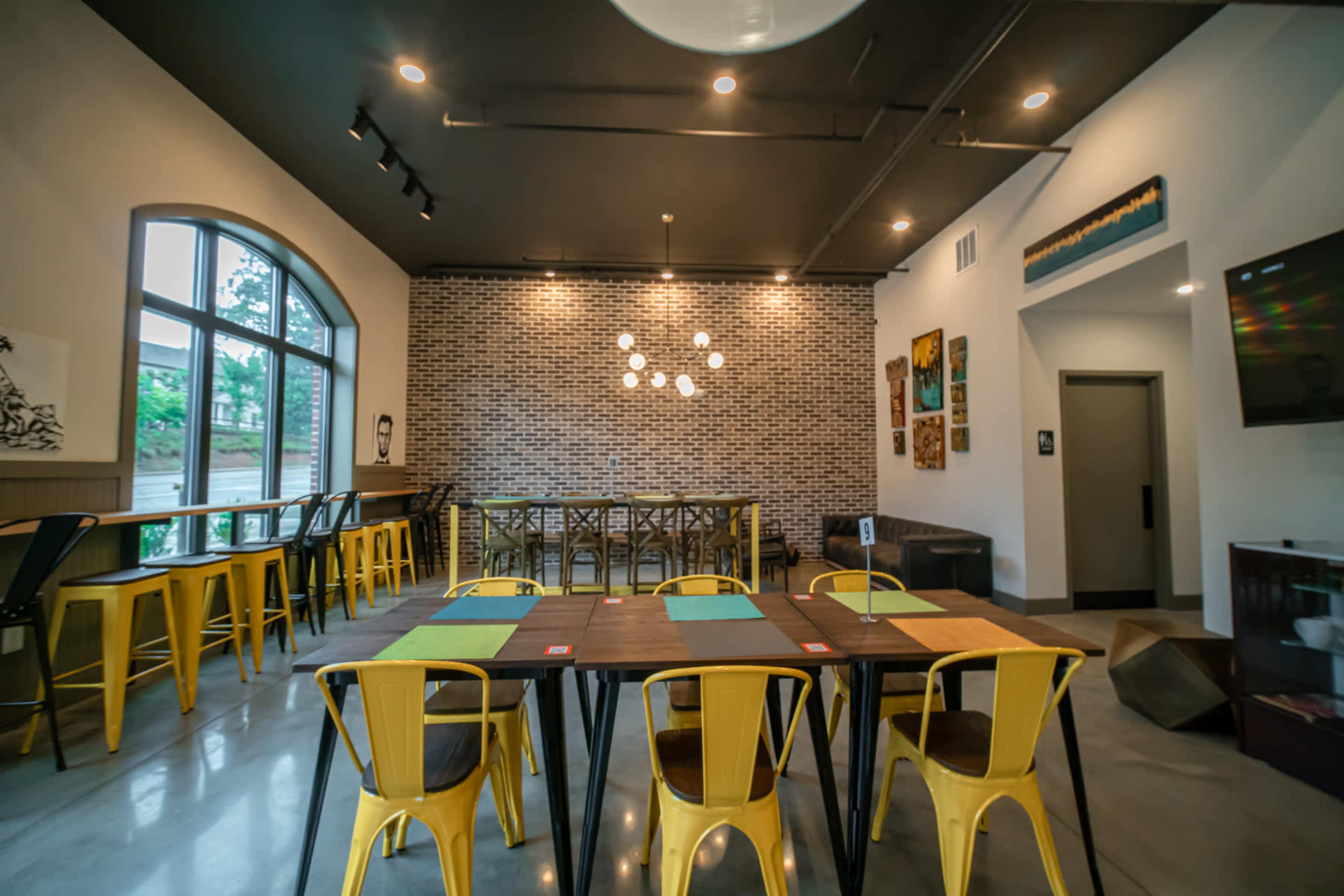 A modern dining space featuring yellow chairs, a large wooden table, and a brick accent wall with pendant lights overhead.