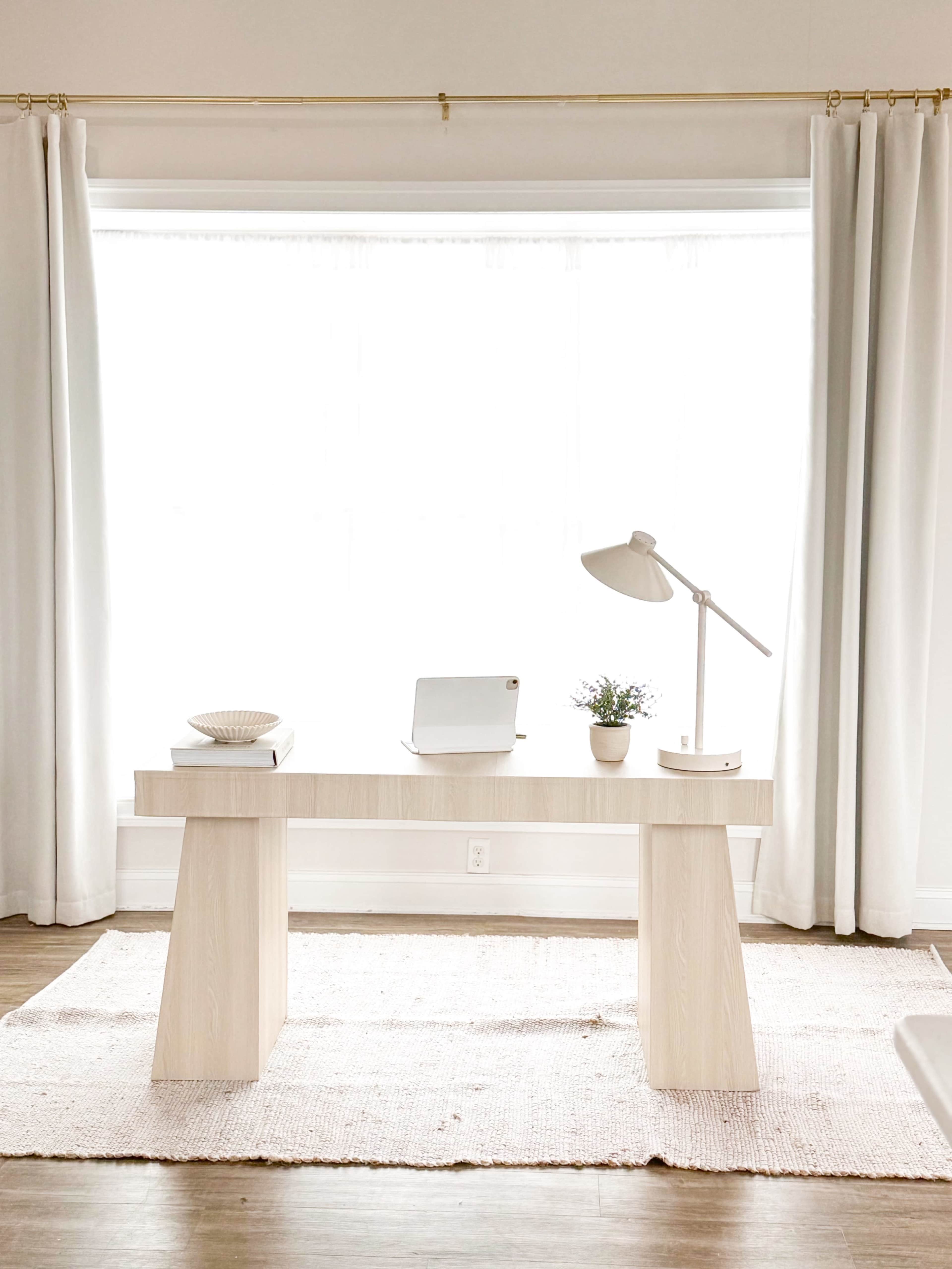 A minimalistic workspace features a wooden desk with a lamp, a small plant, and a laptop, set in front of large windows with white curtains.