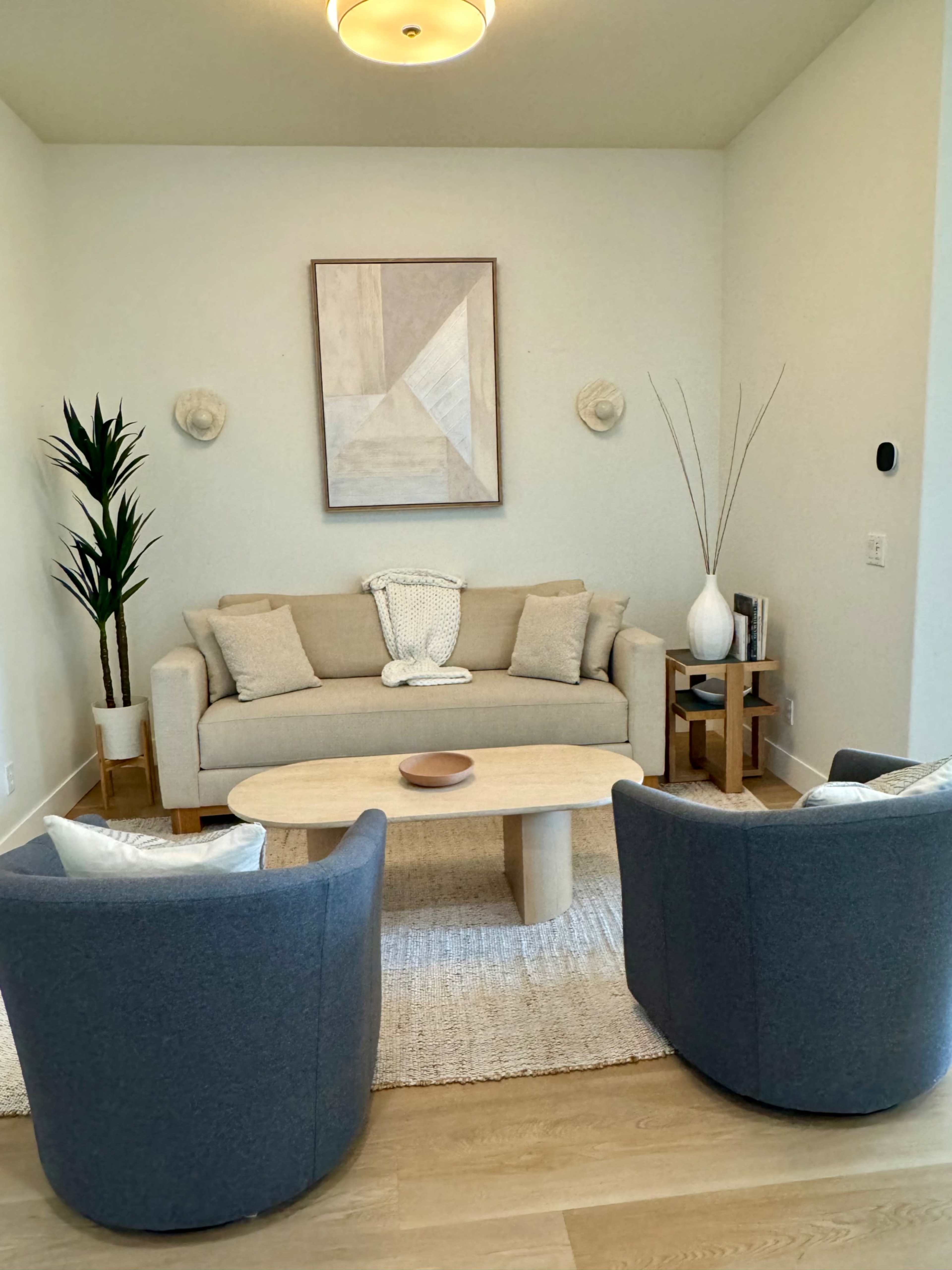 A modern living room featuring a light-colored sofa, two gray chairs, a round wooden coffee table, and decorative plants against a neutral wall.