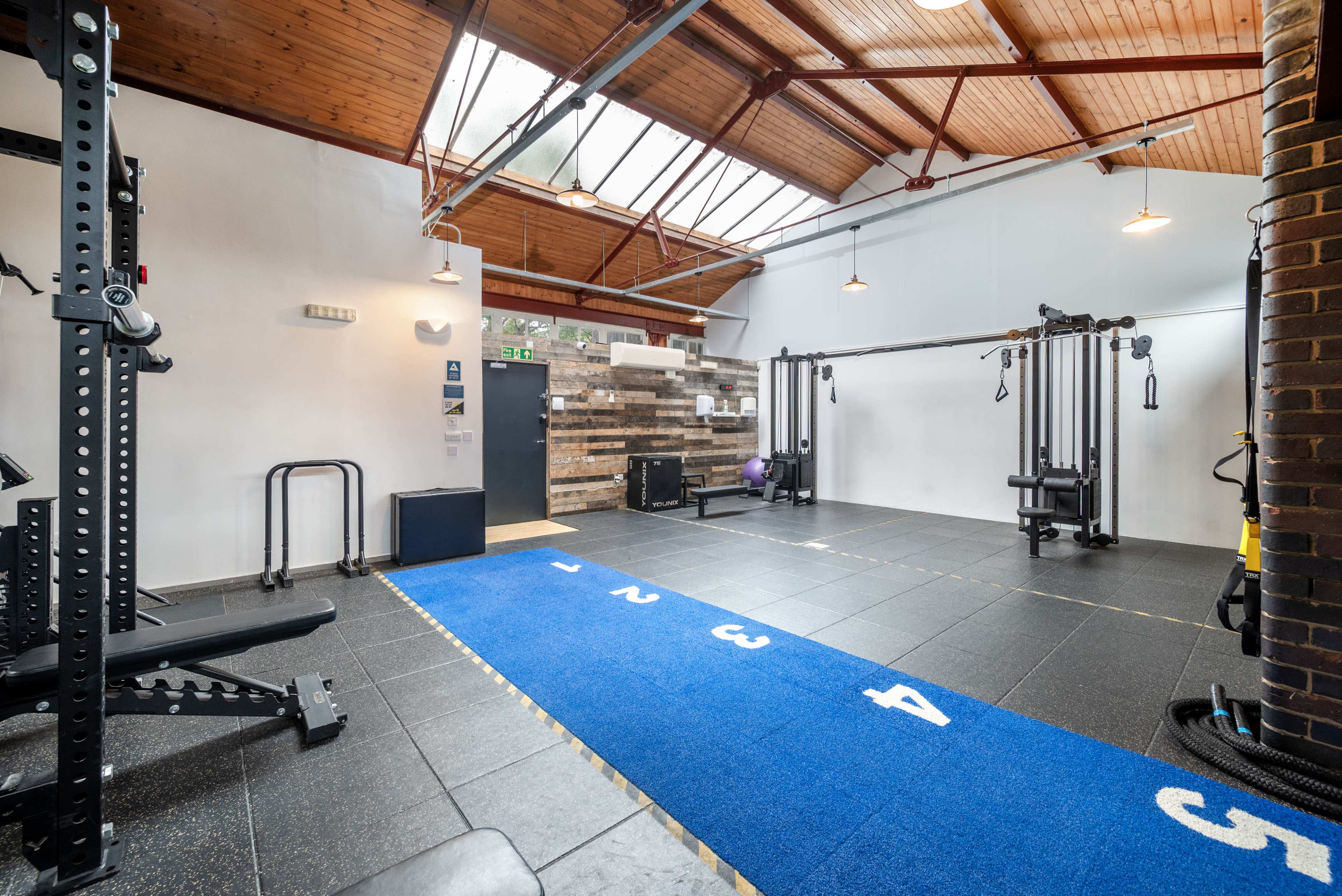 The image shows a modern gym with various exercise equipment arranged on a black tiled floor, and a blue numbered mat running down the center.