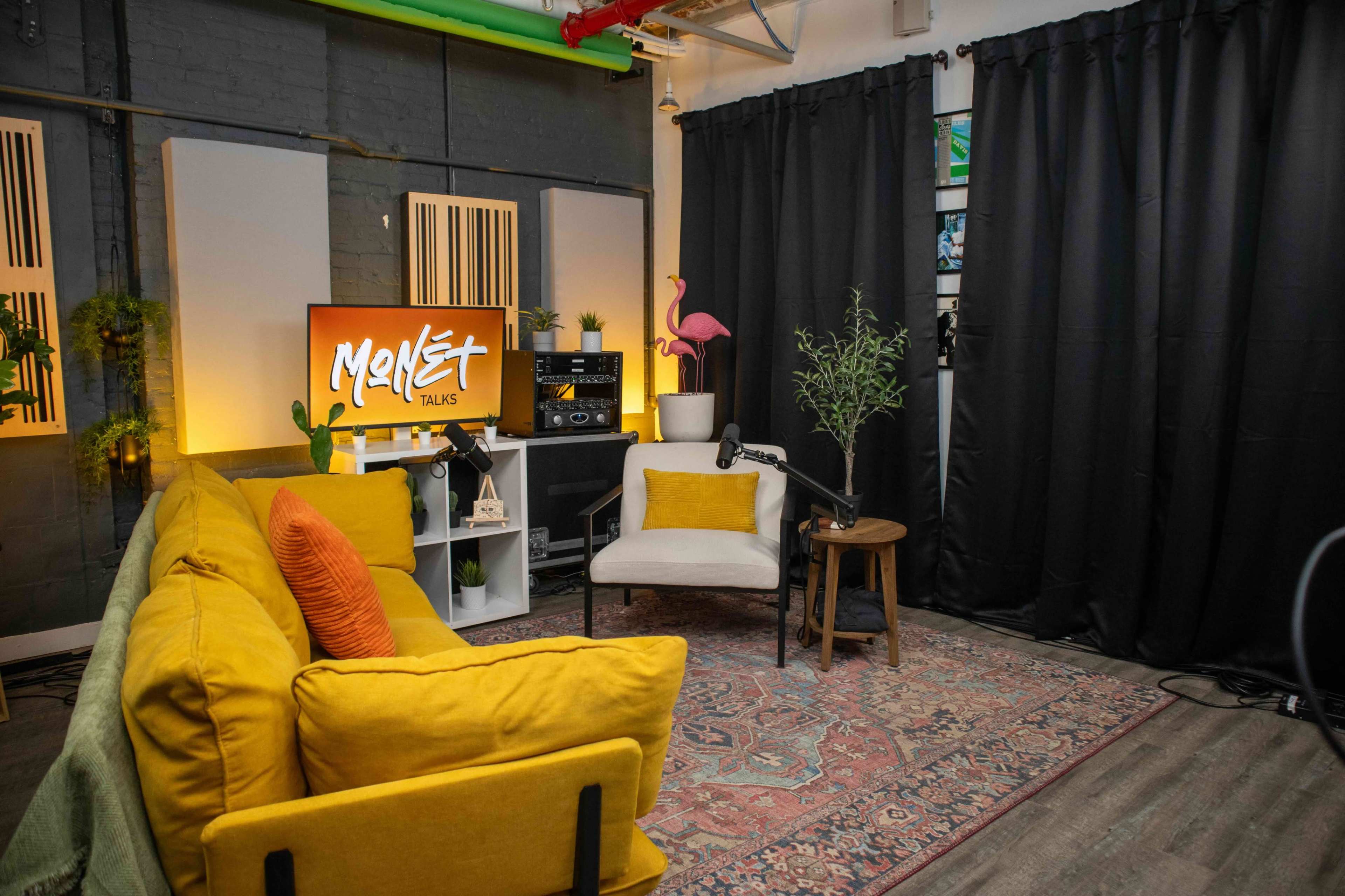 The image shows a cozy room with a yellow sofa, a light-colored chair, and a television displaying "Monset Talks," surrounded by plants and black curtains.