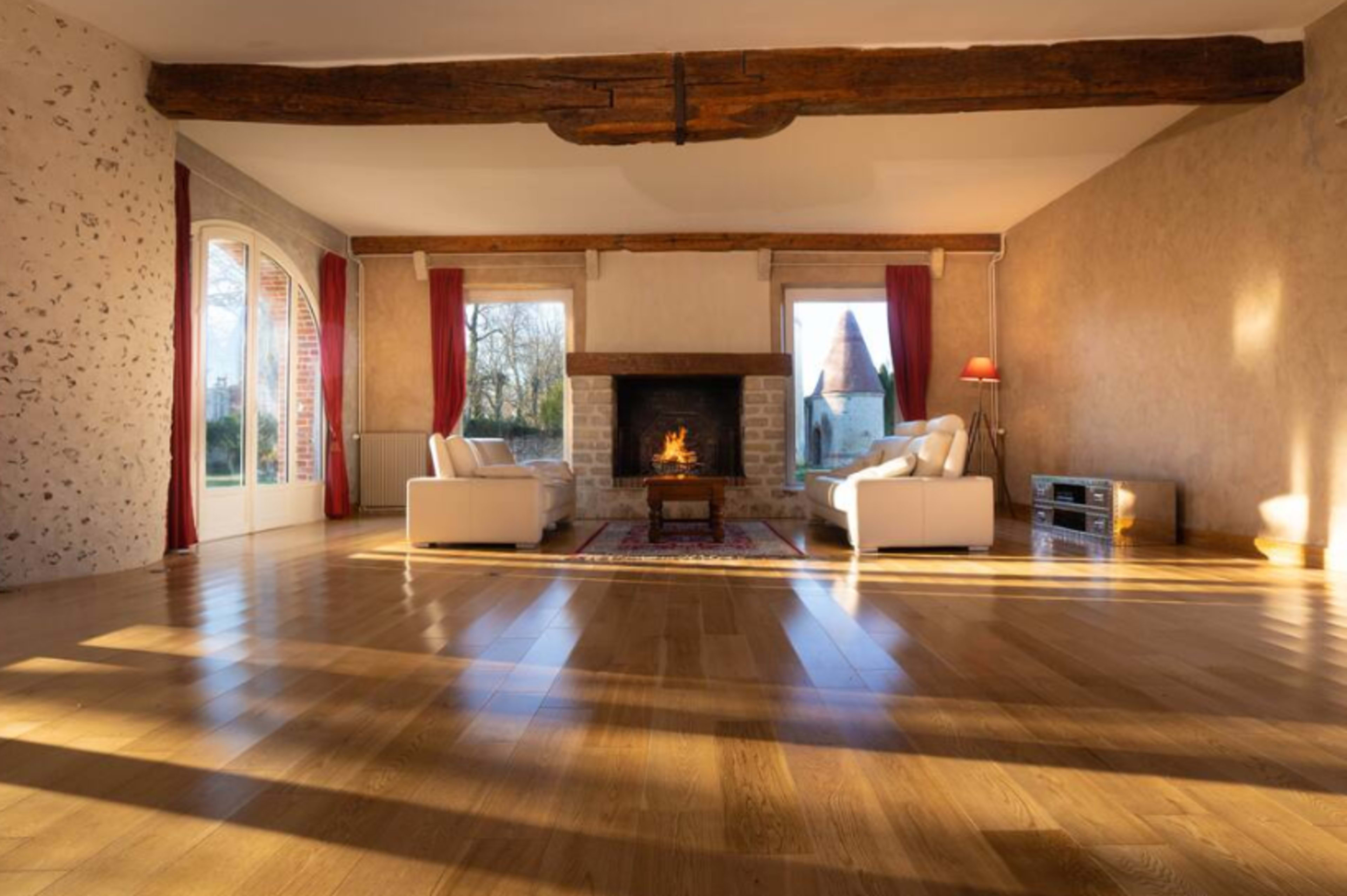 A spacious living room features two white sofas near a stone fireplace, with large windows allowing natural light to fill the space.