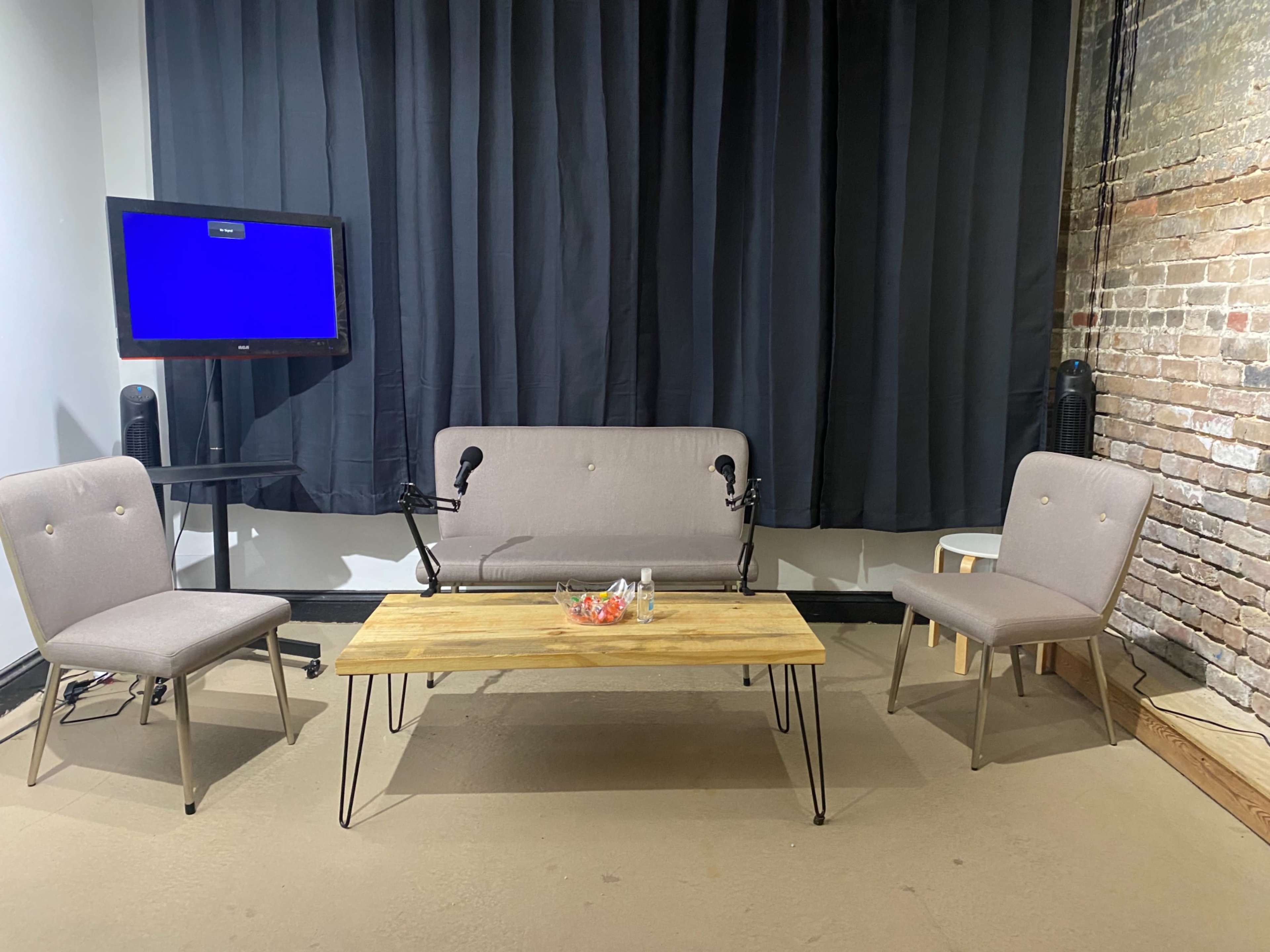 A small interview or discussion set-up with a gray sofa, two chairs, a wooden table with decorative items, and a television screen in front of a dark curtain and a brick wall.