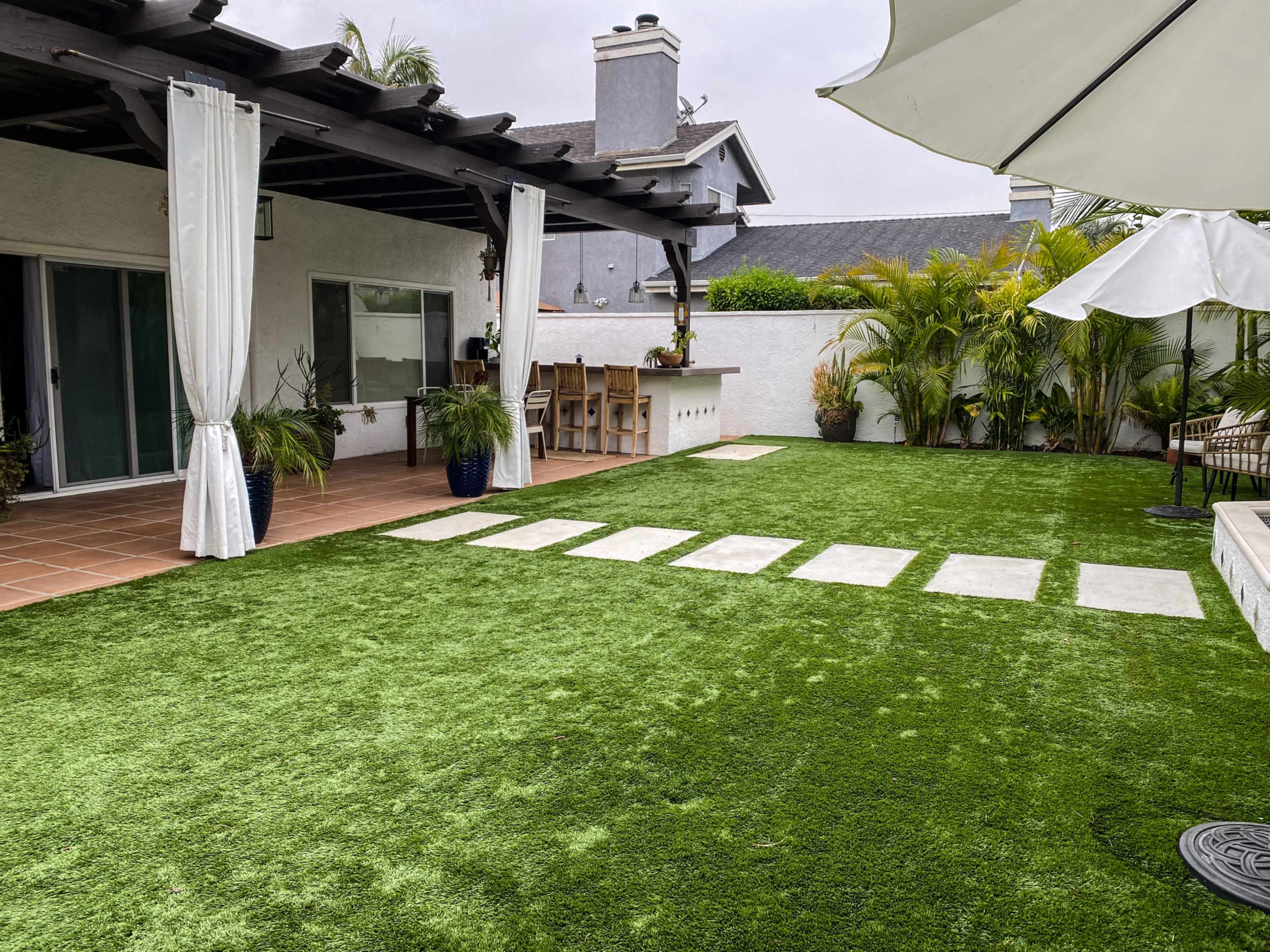 A backyard with artificial grass, a bar area, and a lounge space surrounded by greenery and a covered patio.