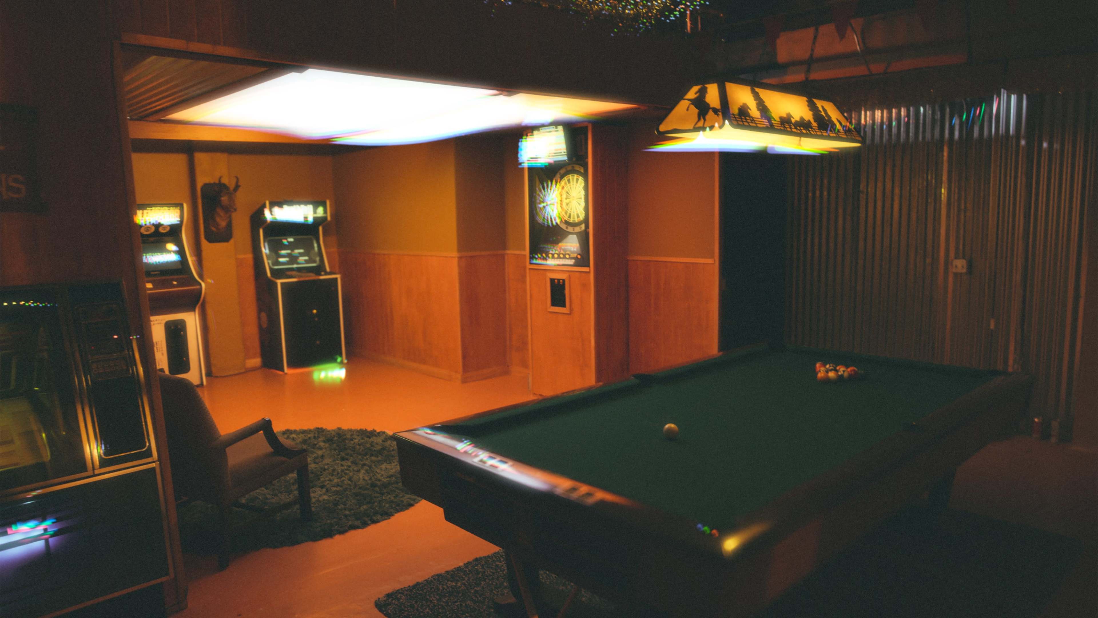The image depicts a dimly lit game room featuring a billiards table, arcade machines, and a dartboard.
