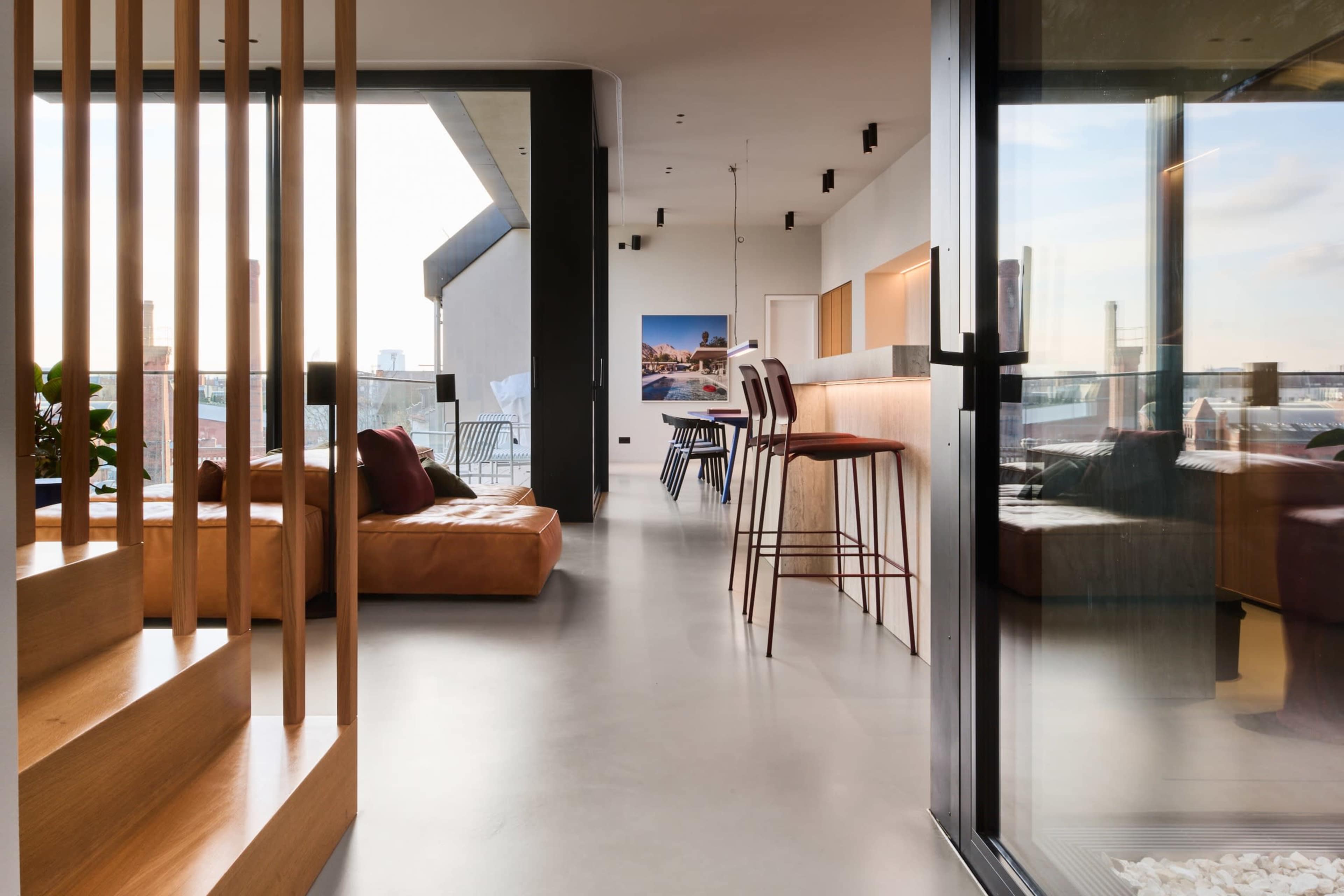 The image shows a modern interior space featuring a staircase, a leather sofa, and a bar with high stools, all illuminated by natural light from large windows.