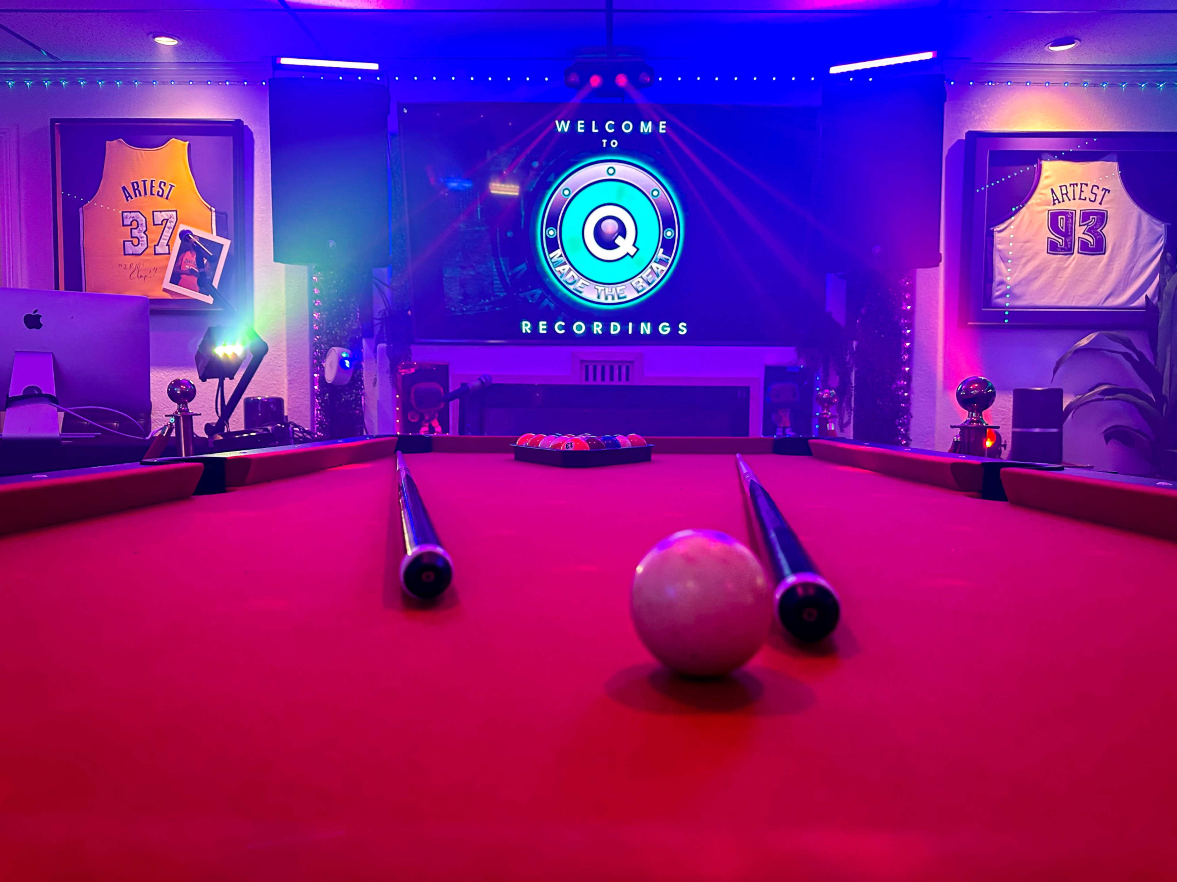 A pool table with a white ball in the foreground is centered in a room featuring colorful lighting and framed jerseys on the walls.