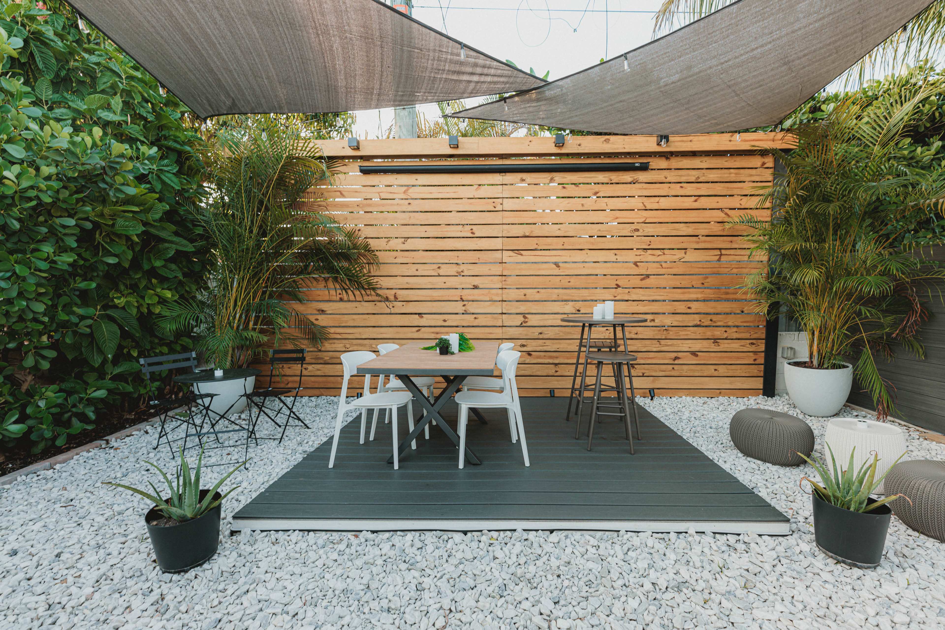 The image depicts a modern outdoor patio area featuring a wooden slatted backdrop, a dark deck with a table and chairs, and various plants arranged in decorative pots.