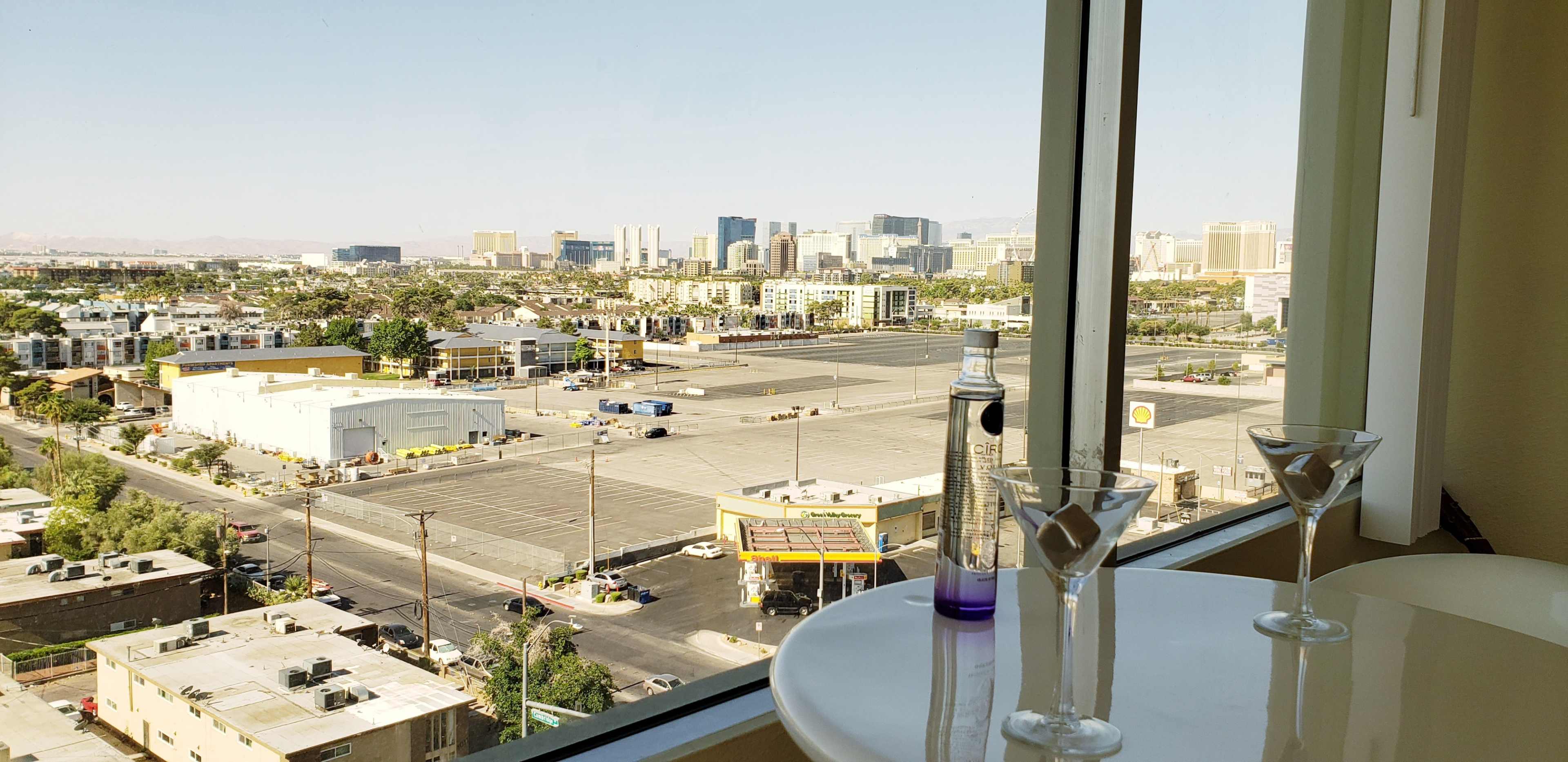A view of a city skyline with a parking lot and buildings from a high-rise window, accompanied by a table featuring two martini glasses and a bottle.
