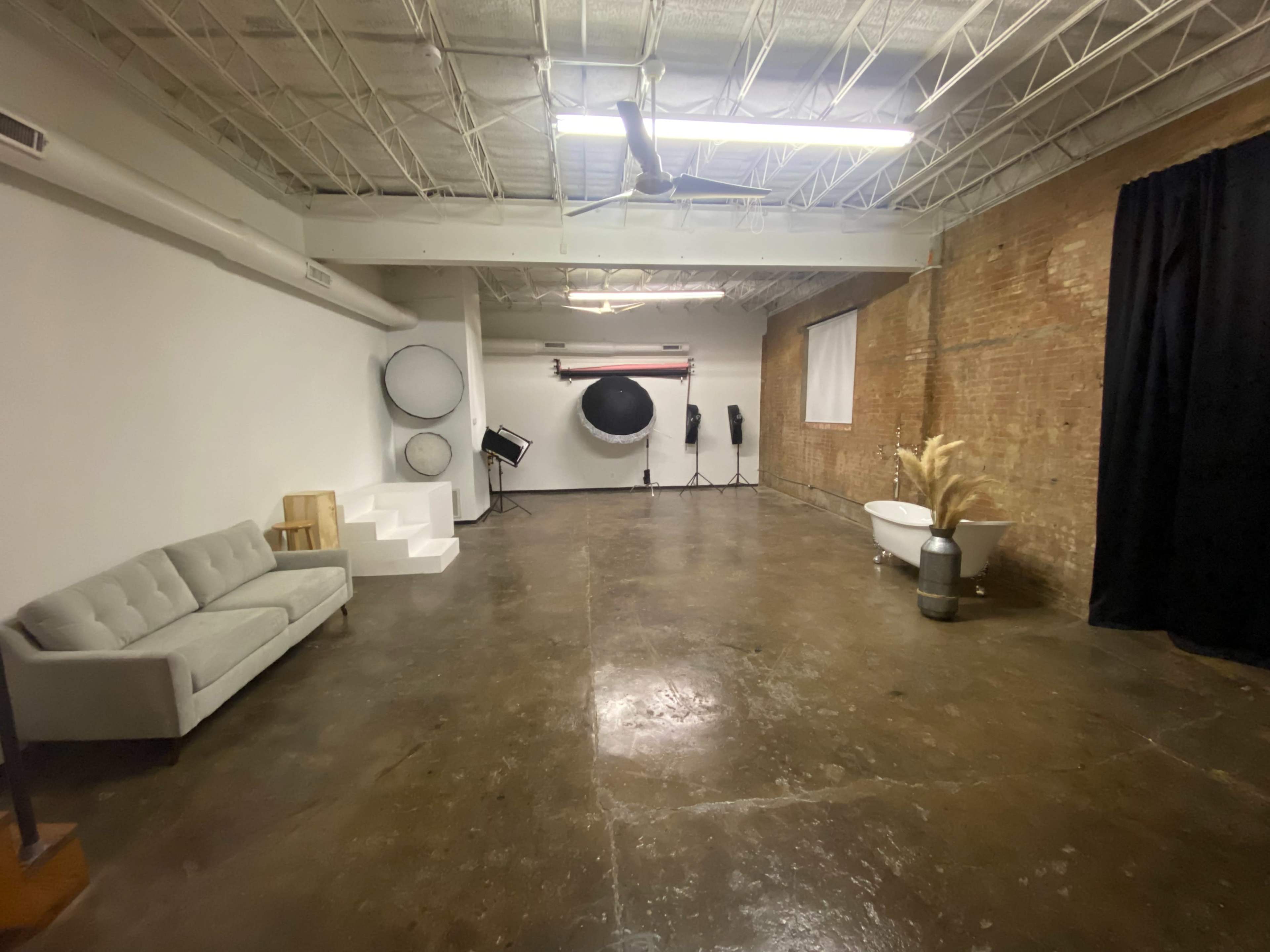 A spacious, industrial-style studio featuring a gray couch, a circular backdrop, and various photography equipment against a brick wall.