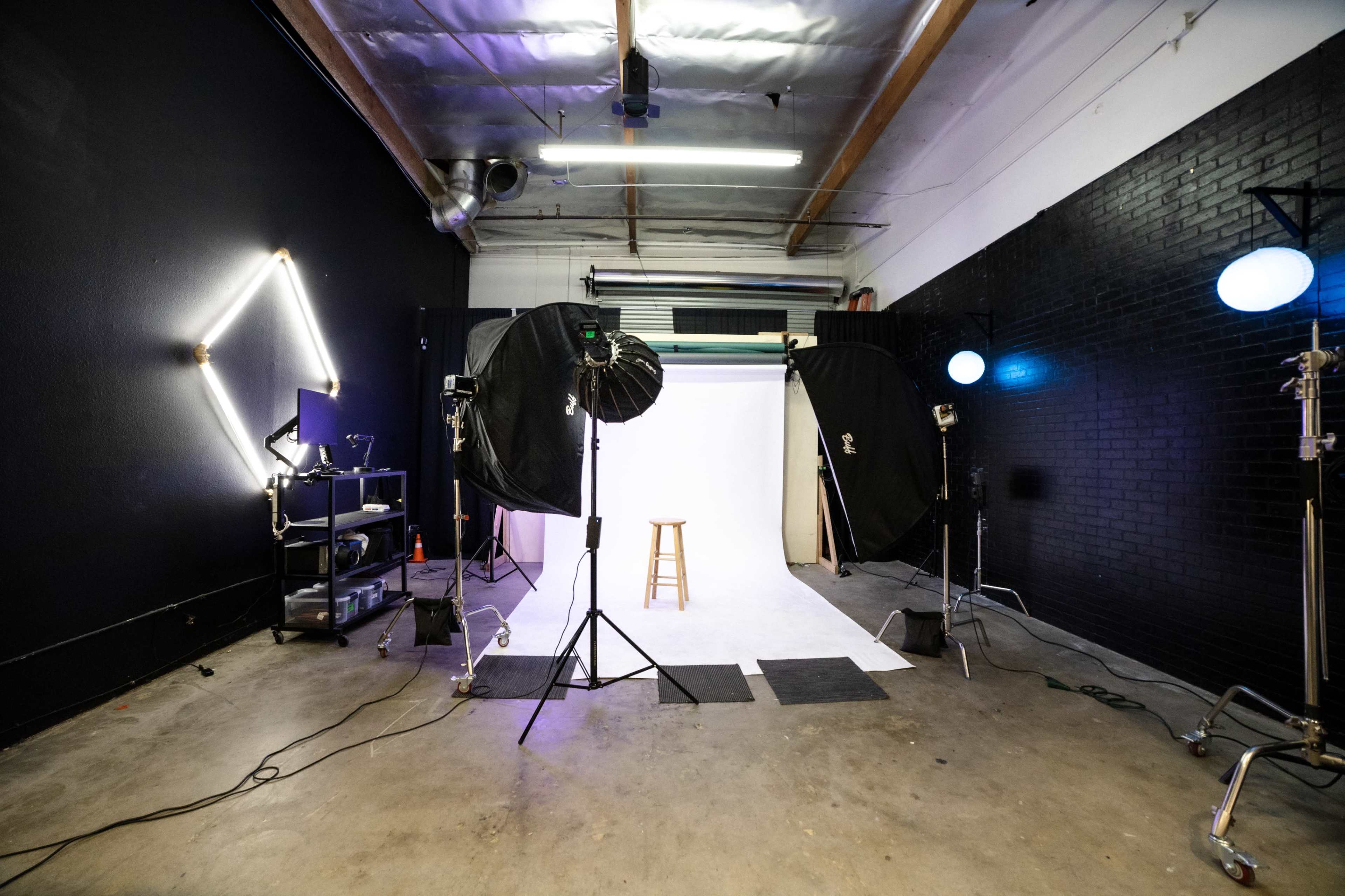 The image shows a photography studio setup with a white backdrop, lighting equipment, and a wooden stool in the center.