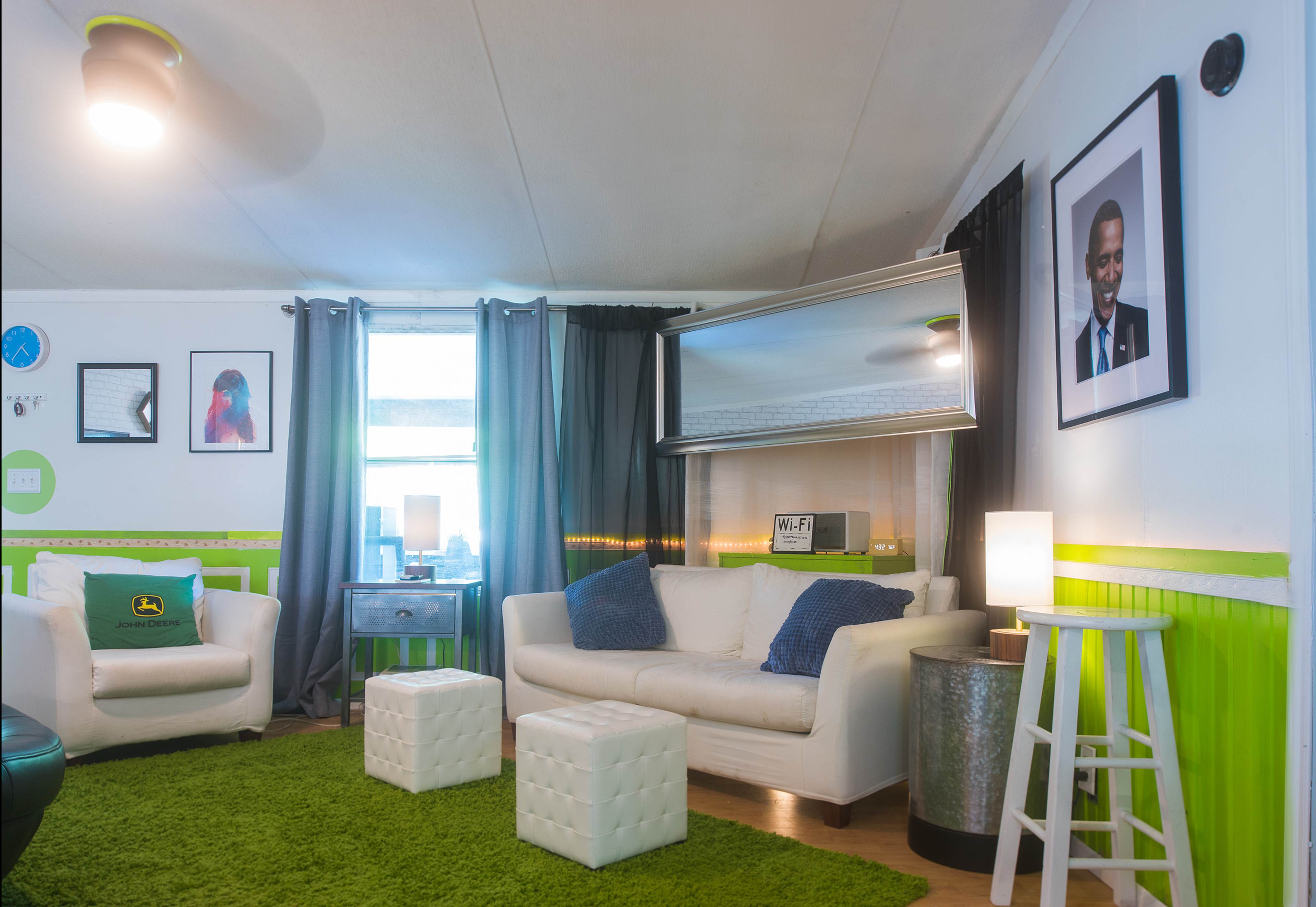 The image shows a cozy living room with white sofas, green carpeting, decorative framed artwork on the walls, and a large mirror.
