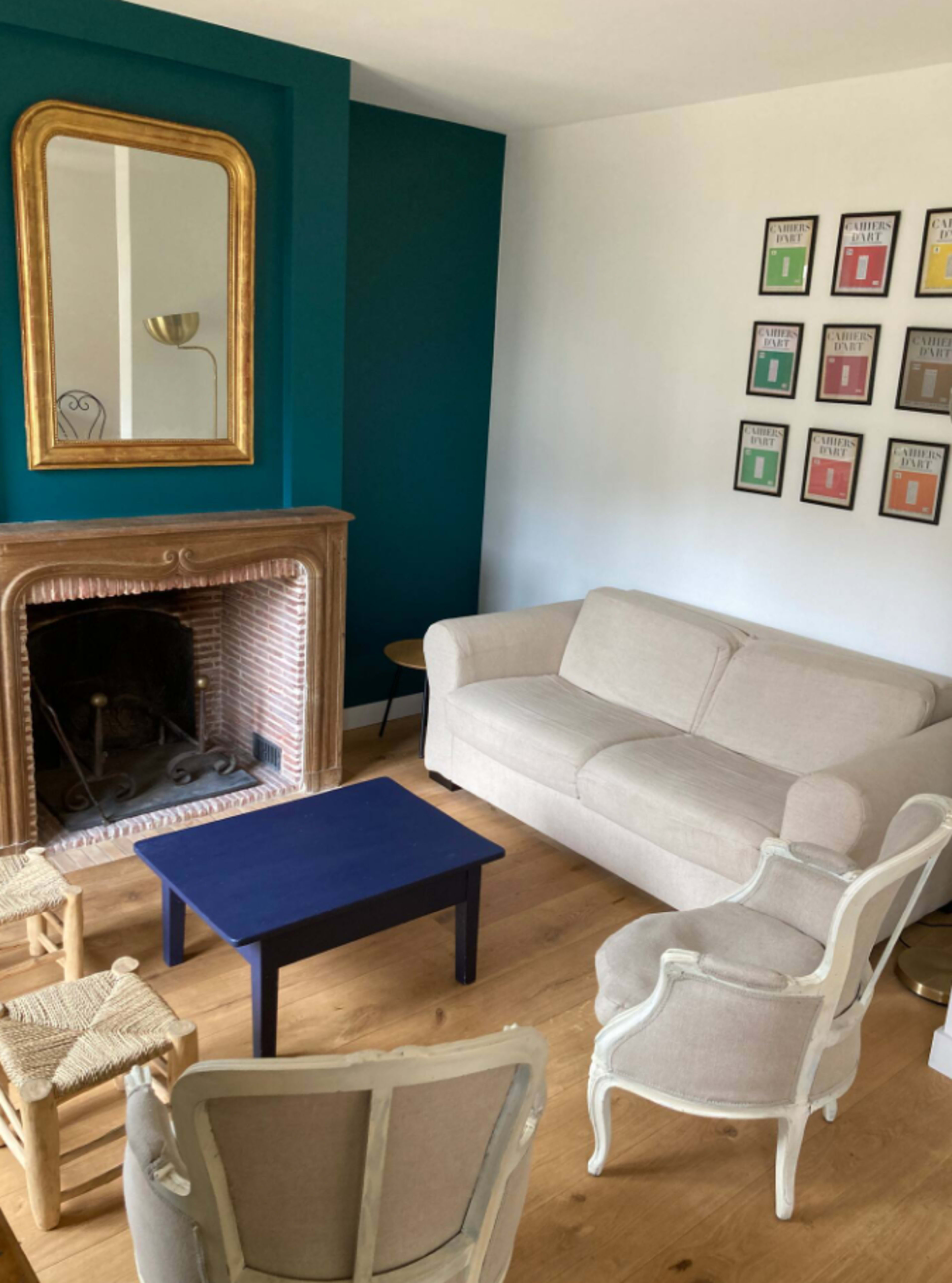 The room features a beige sofa, two white armchairs, a blue coffee table, and a fireplace, with a mirror and a series of framed prints on the walls.