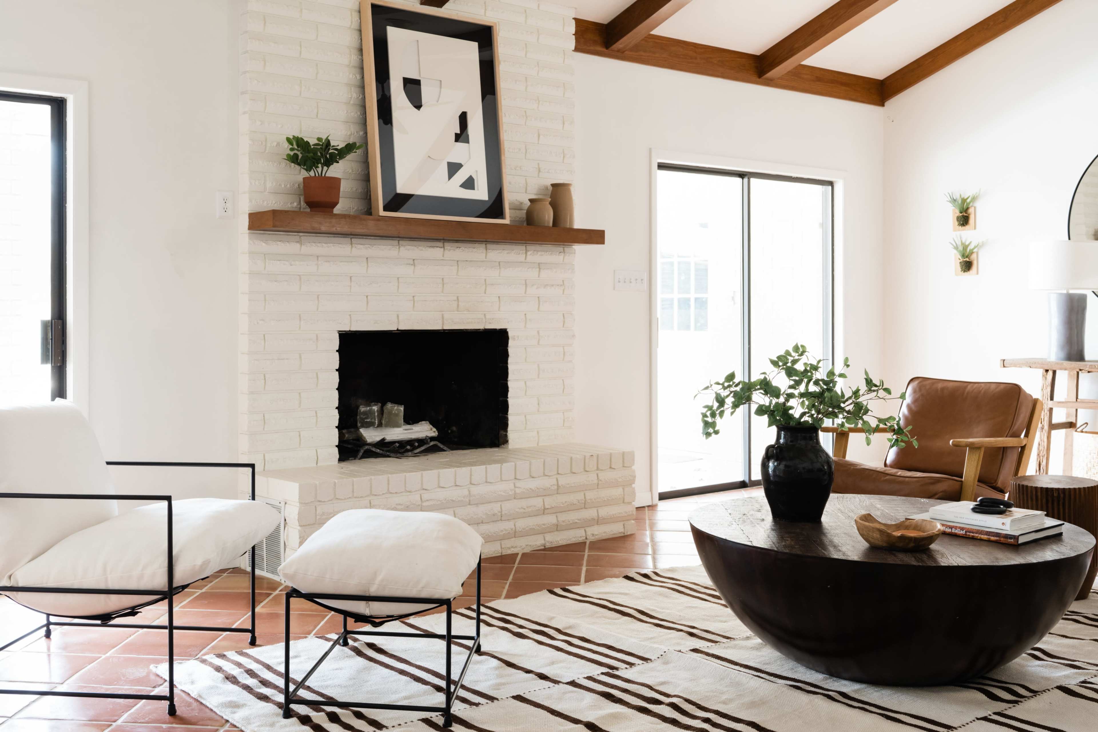 A living room featuring a white brick fireplace, a dark round coffee table, two chairs, and large windows providing natural light.