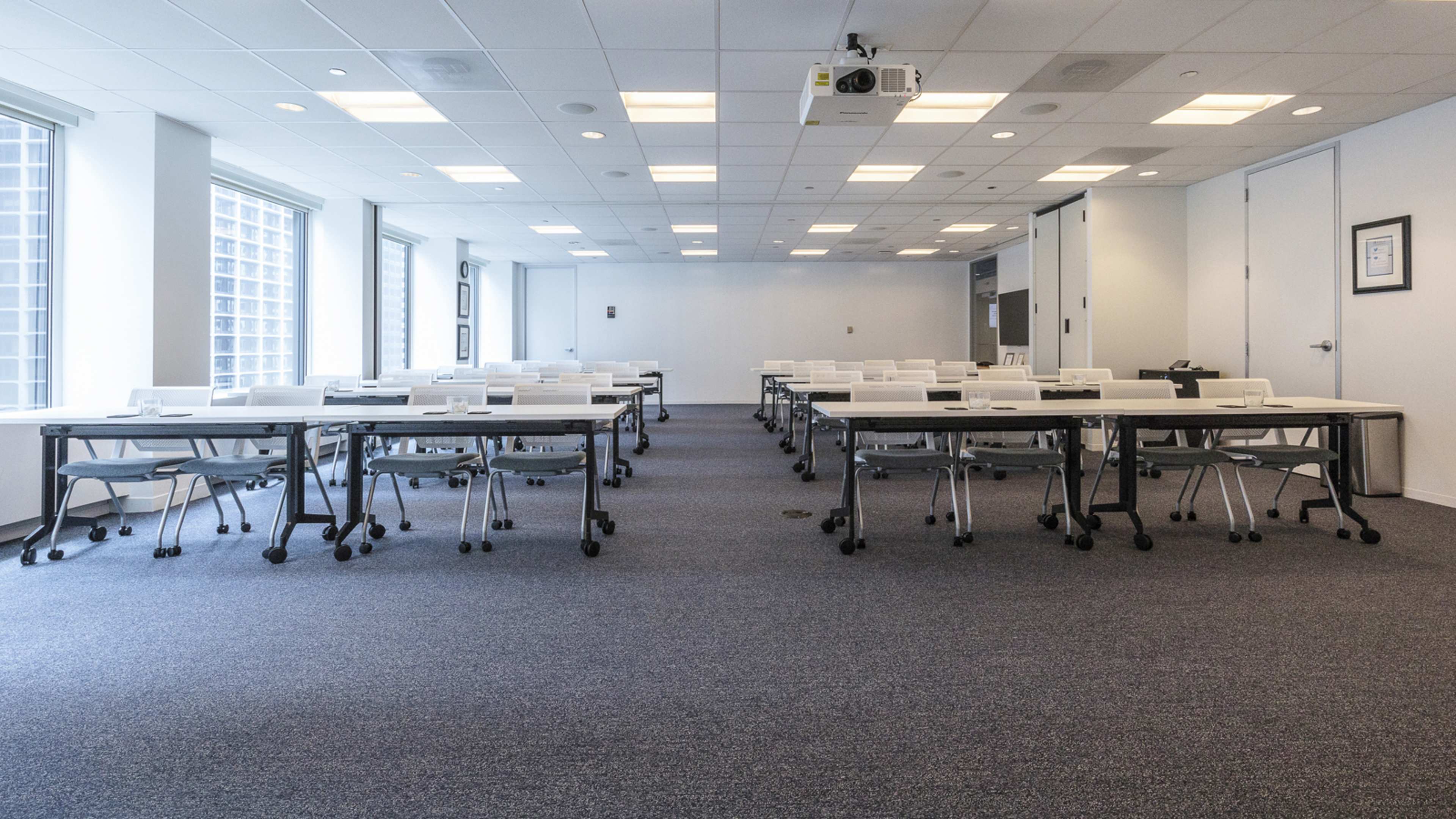 A conference room is set up with multiple tables and chairs arranged in rows, featuring a wall-mounted projector and windows providing natural light.