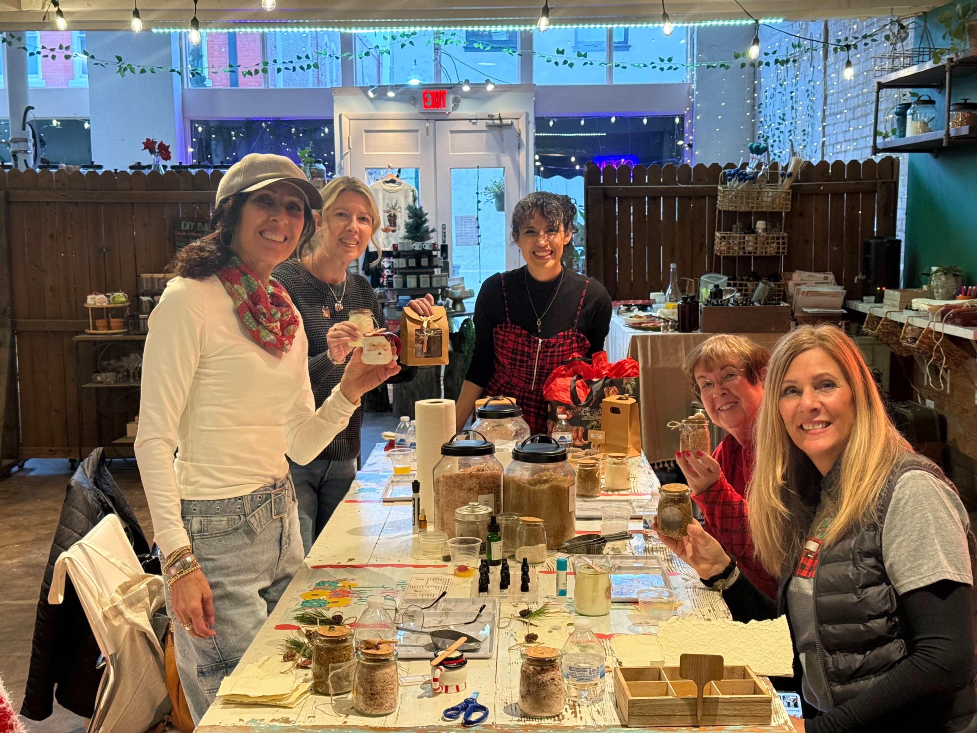 A group of five women stands around a table filled with jars and crafting materials in a brightly lit shop.