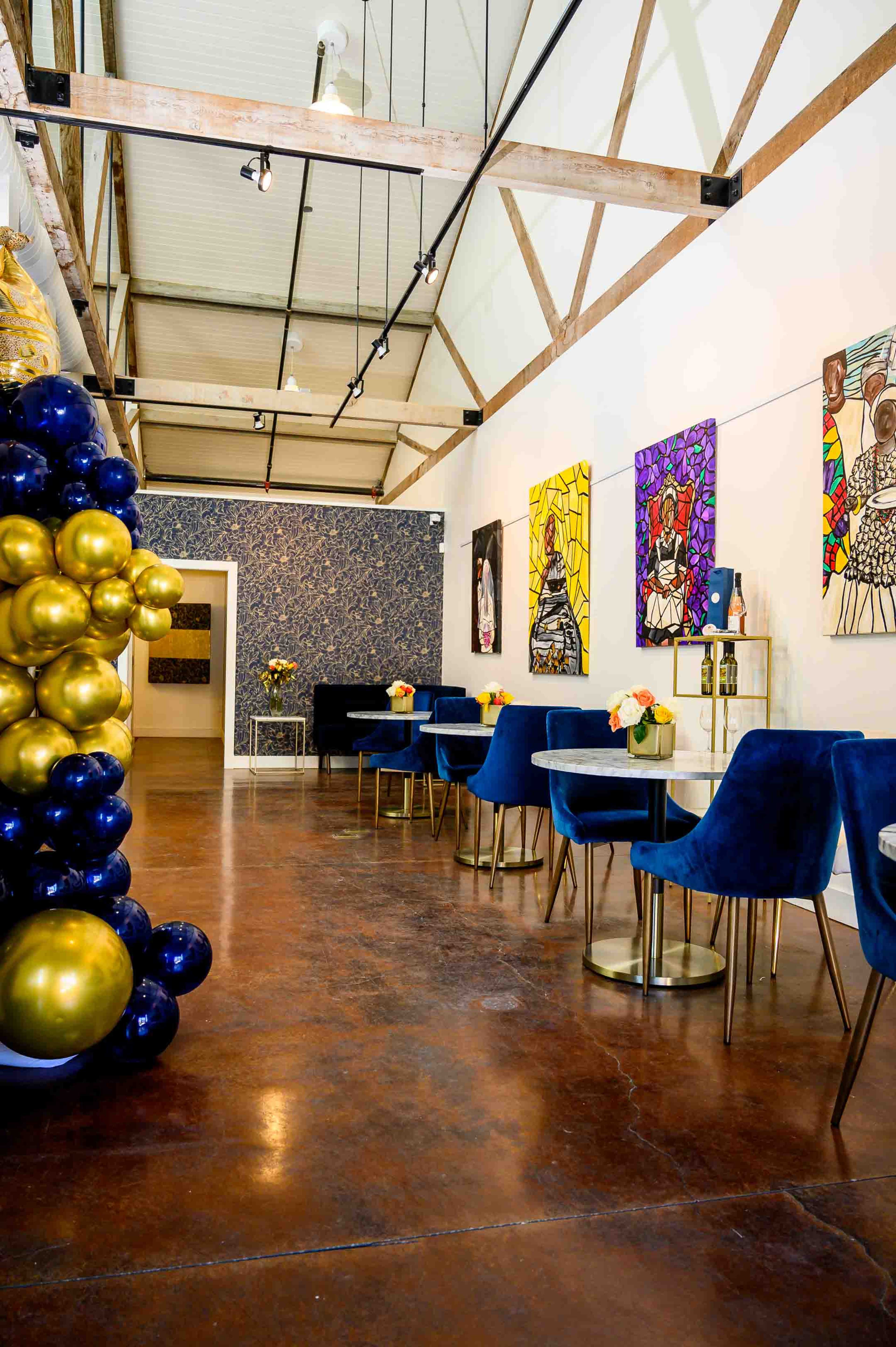 The image shows a contemporary interior of a cafe featuring blue velvet chairs, gold and navy balloon decorations, and colorful artwork on the walls.