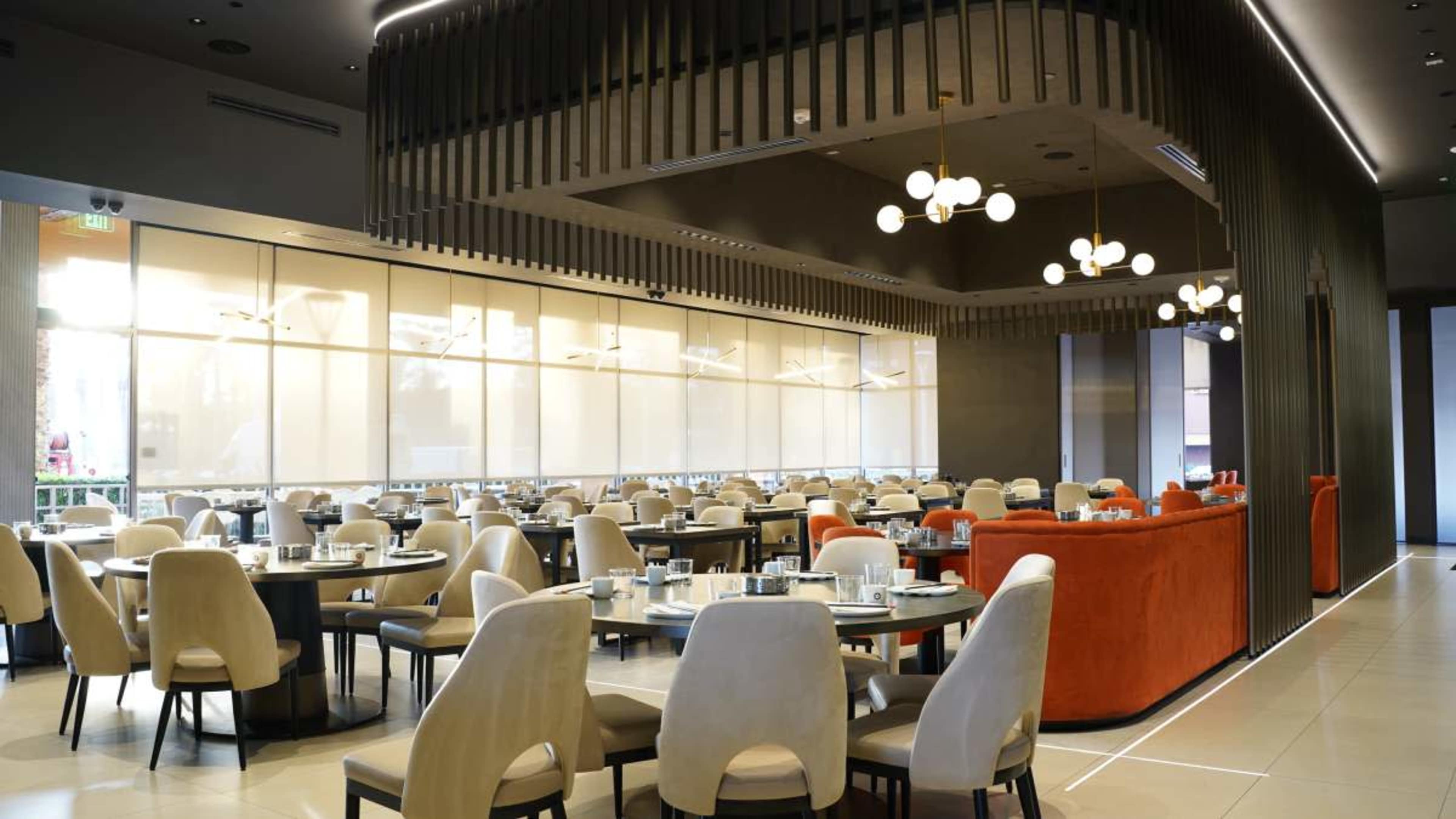The image shows a modern restaurant interior with numerous dining tables and chairs arranged neatly, large windows allowing natural light, and a distinctive ceiling design.