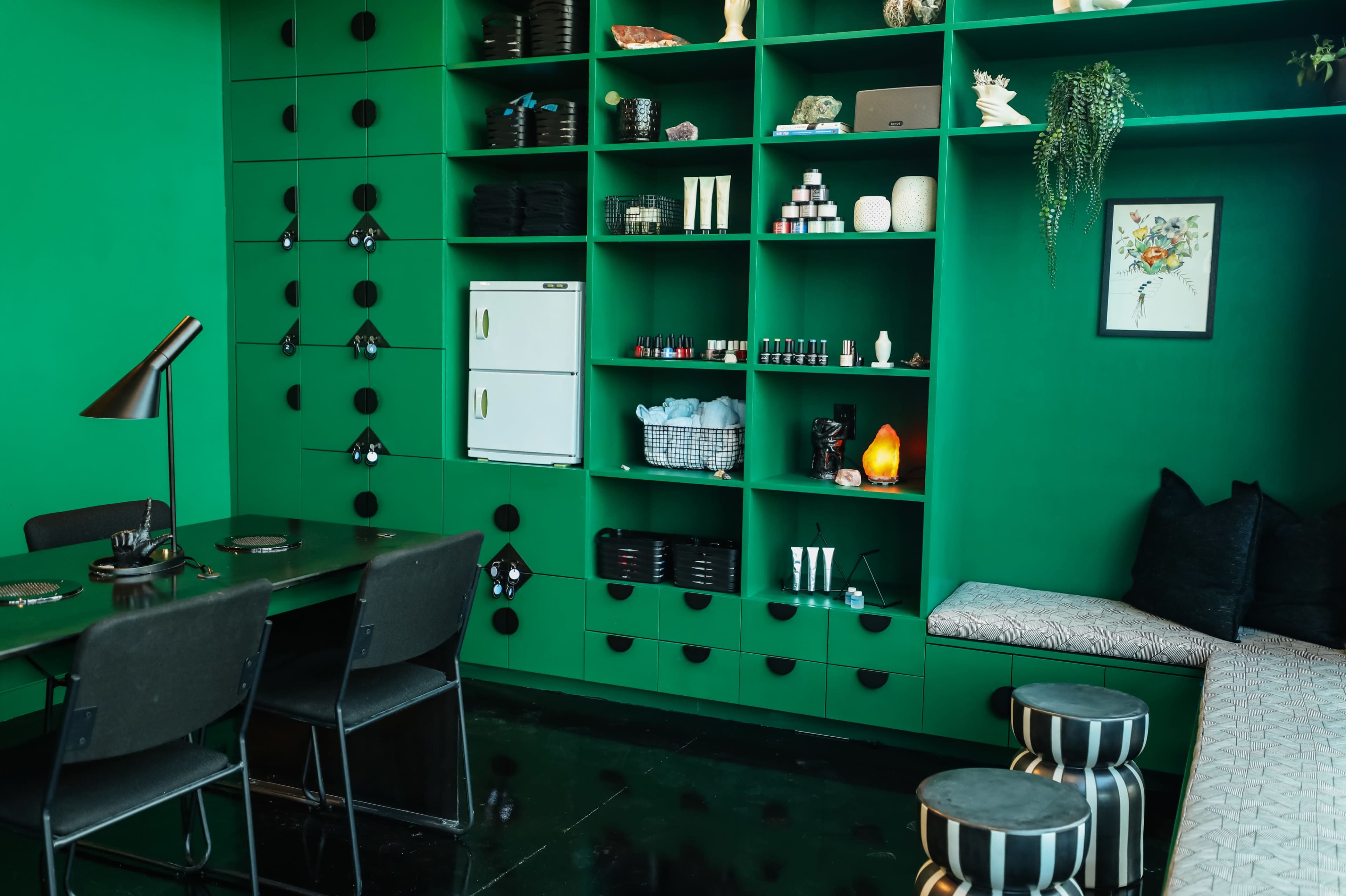 A green-walled room features a shelf filled with assorted items, a small refrigerator, a table with black chairs, and a sofa with decorative cushions.