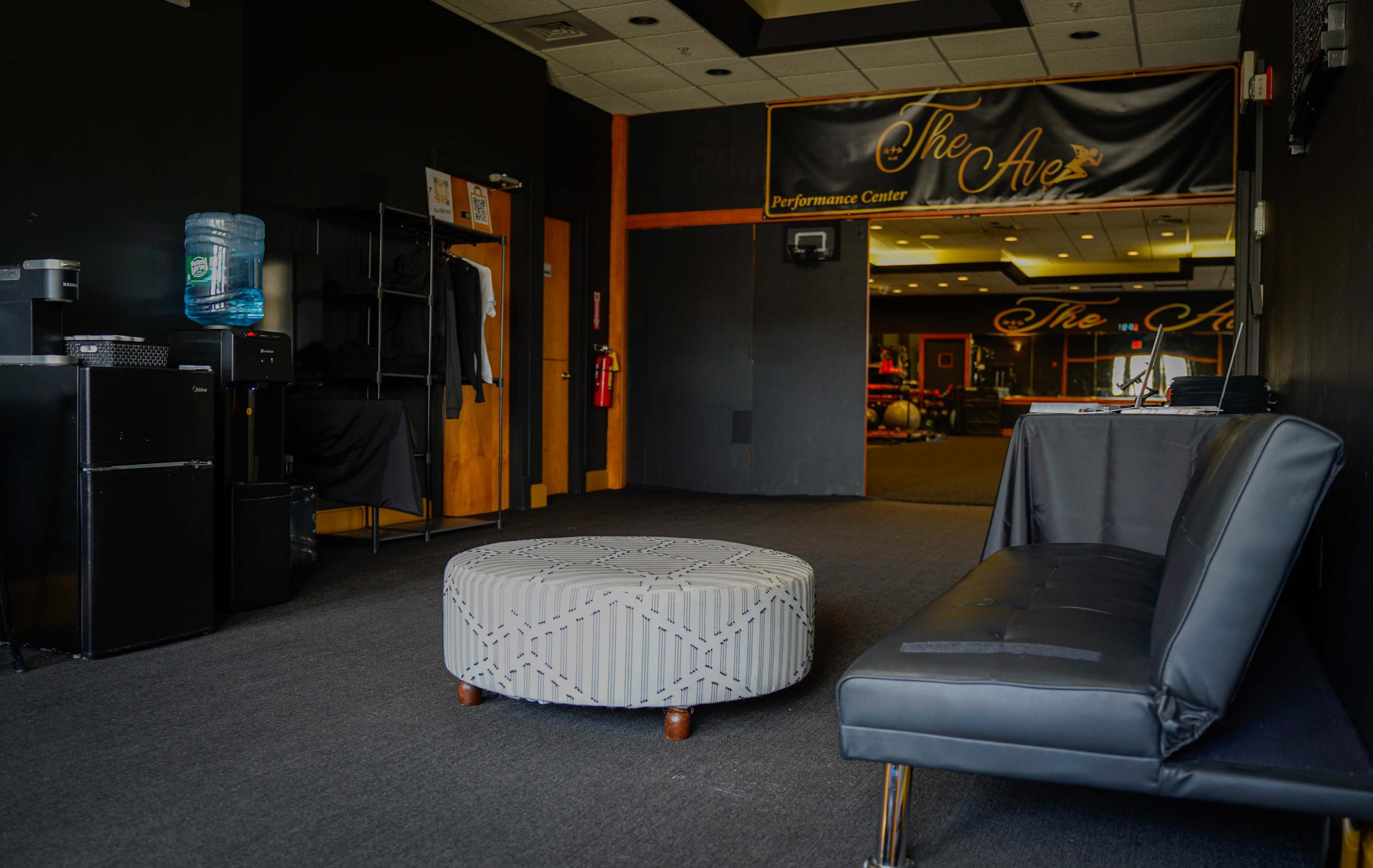 A lounge area with a black sofa and a patterned ottoman, leading to a performance center in the background.