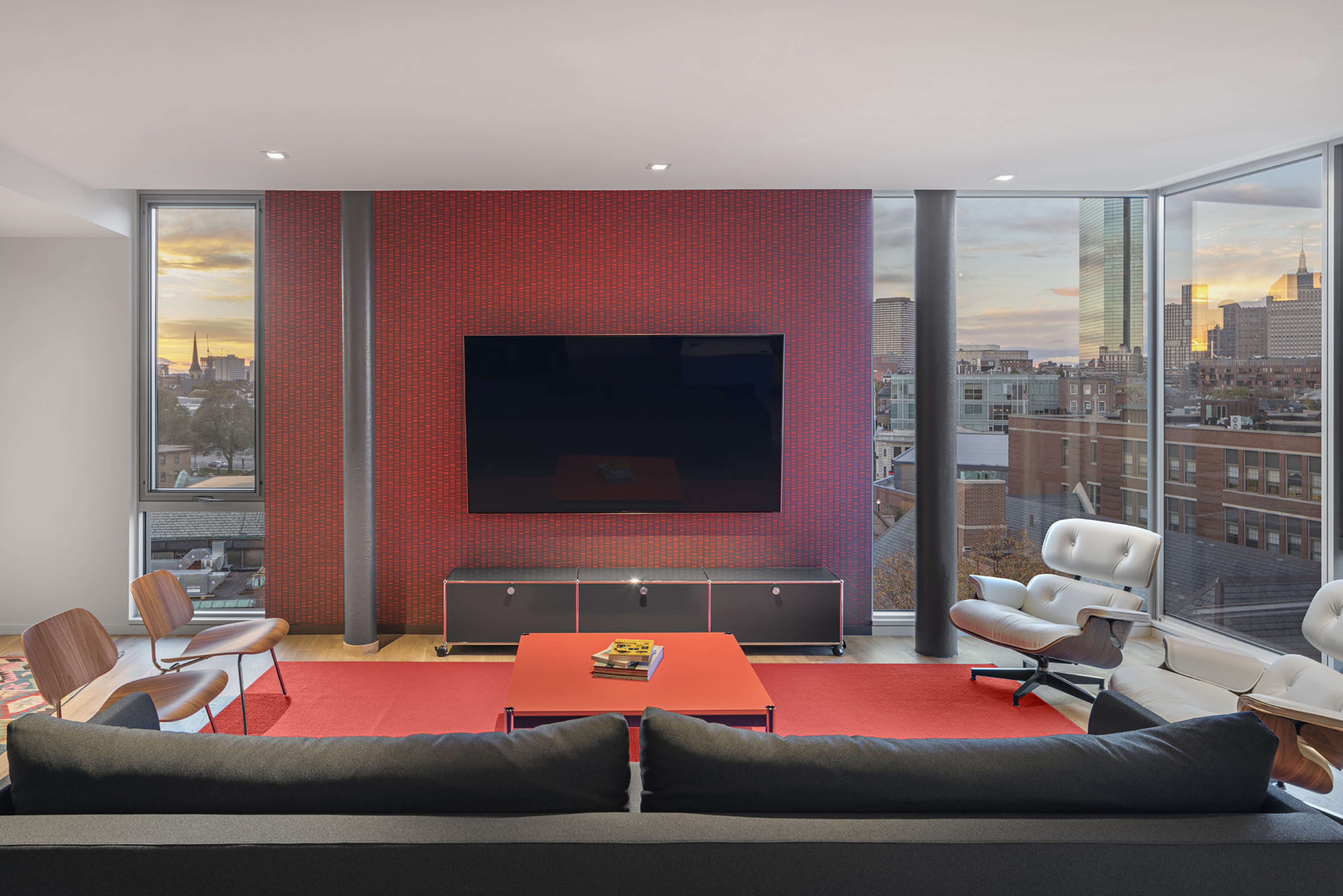 A modern living room features a large television mounted on a red patterned wall, surrounded by contemporary furniture and large windows showcasing an urban view.
