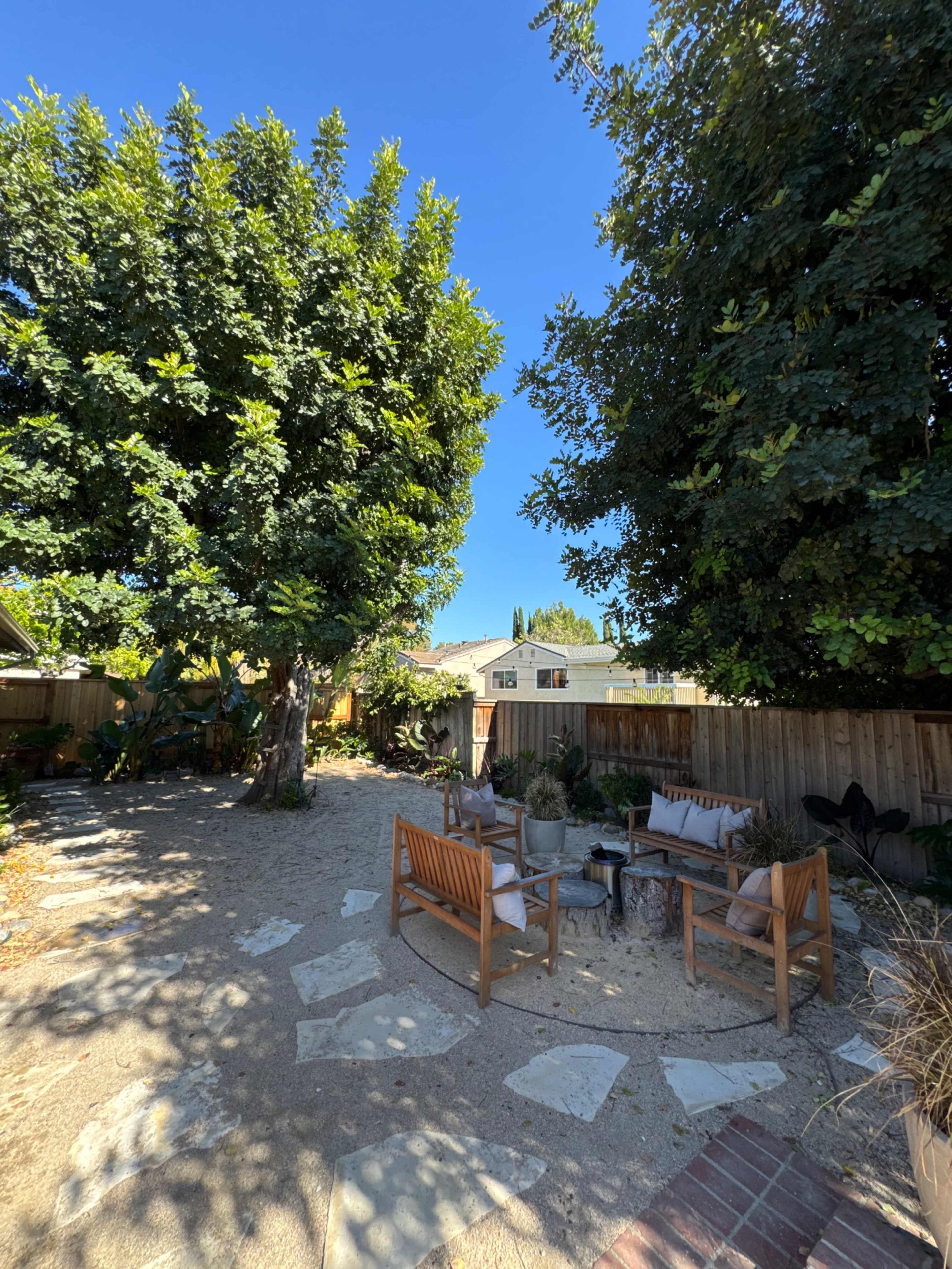 The image depicts a backyard with two large trees, stone pathways, and a seating area furnished with wooden chairs and tables.