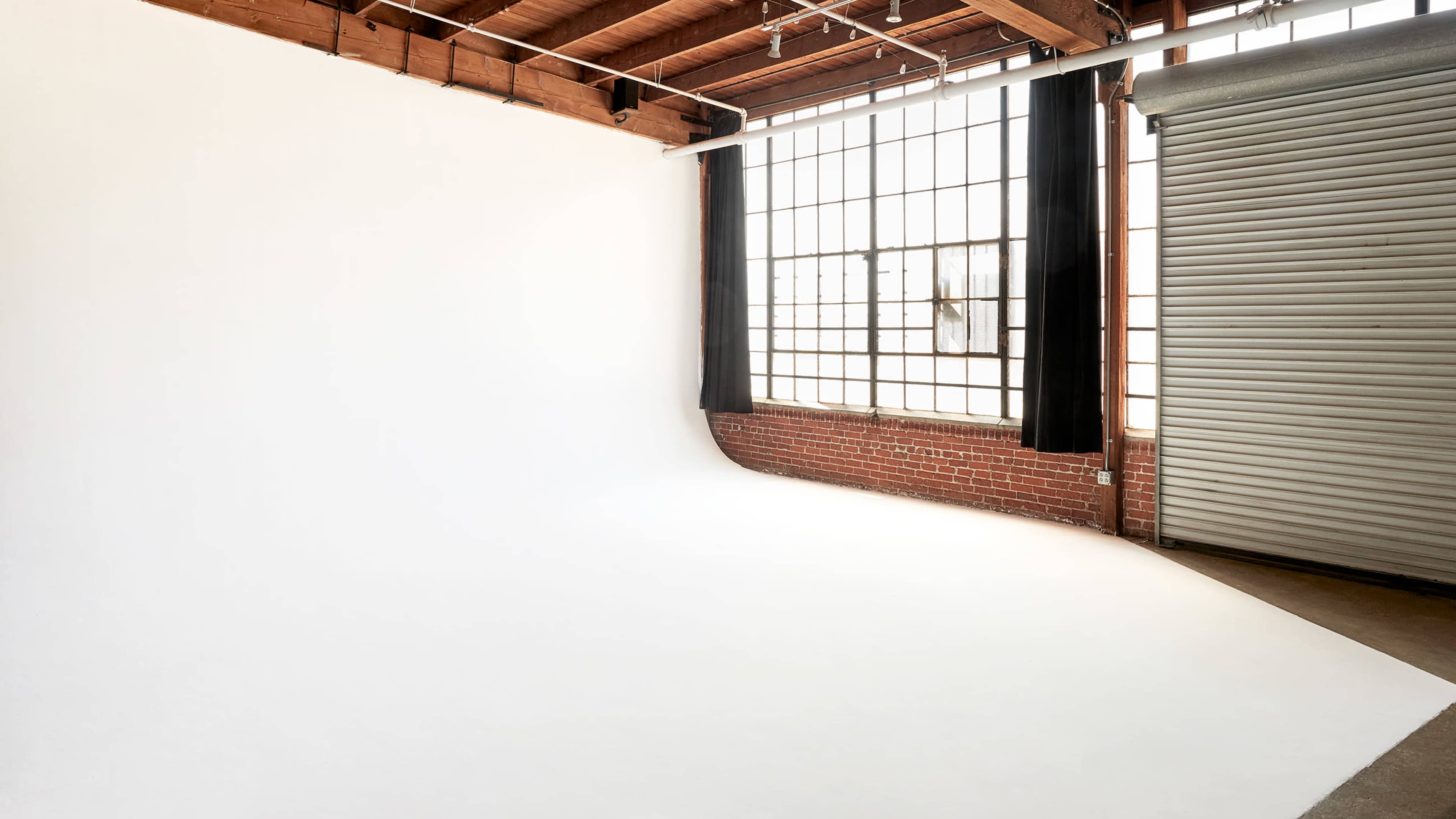 The image shows a spacious photography studio with a white backdrop, large windows, and a brick wall.
