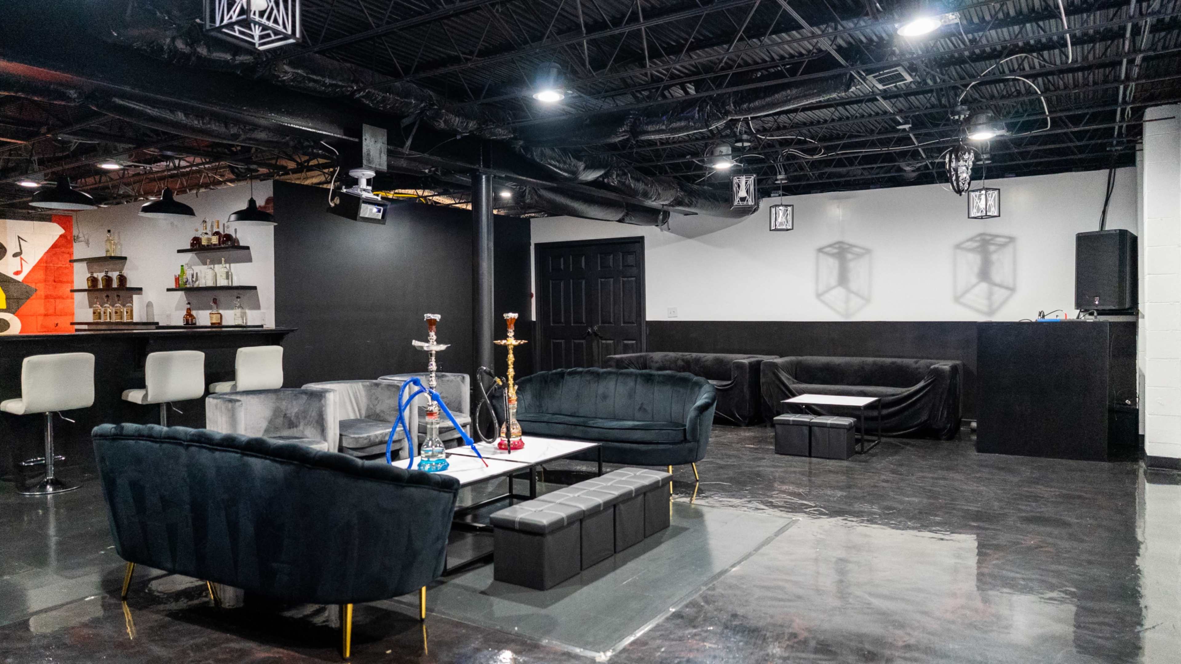 A spacious lounge area featuring dark furniture, a bar with liquor bottles, and a setup for hookahs in a modern interior.