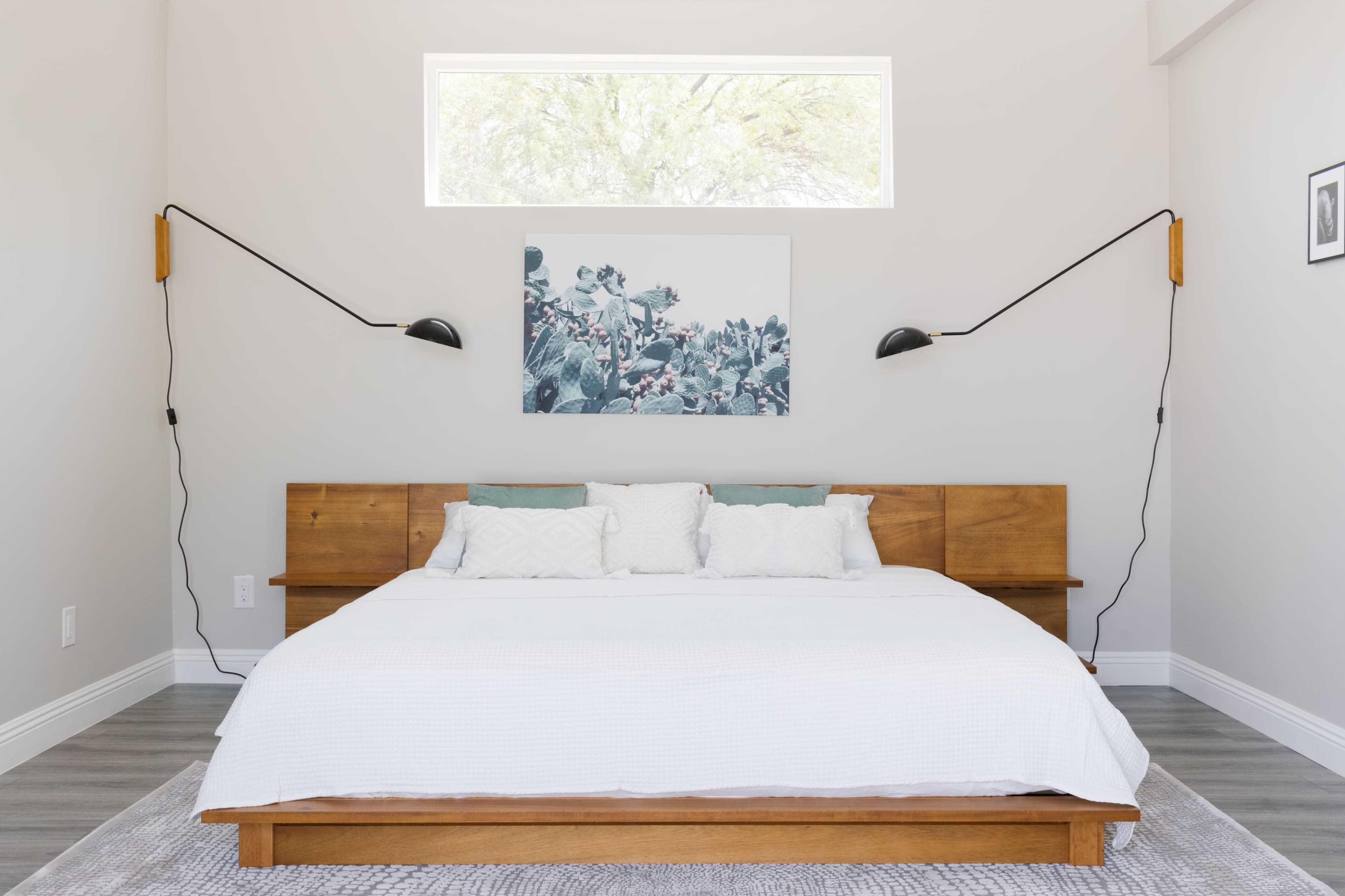 A minimalist bedroom features a wooden bed with white bedding, two mounted black wall sconces, and a large window above a floral artwork.