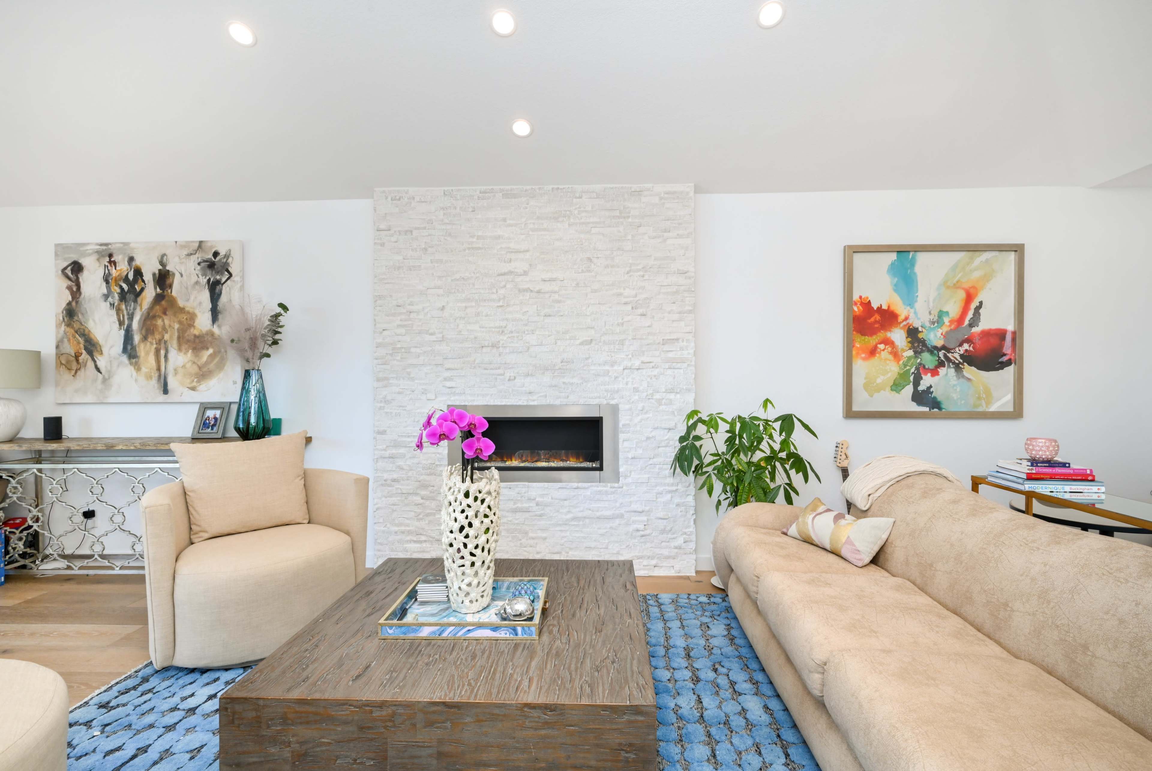 A modern living room with a beige sofa, a wooden coffee table, a decorative vase with purple flowers, and a white stone fireplace.