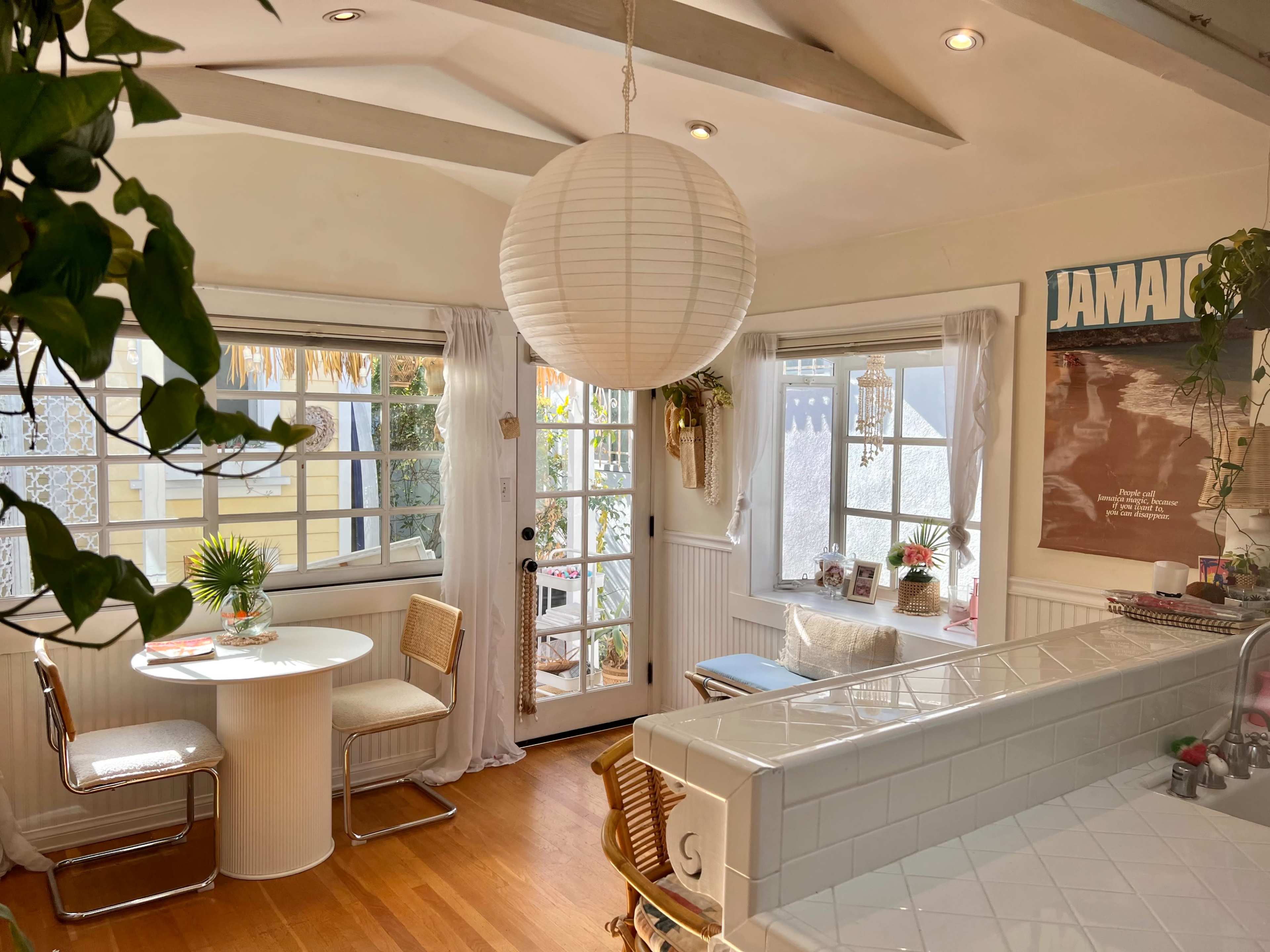A bright dining area features a white round table with two chairs, large windows, and a paper lantern hanging from the ceiling.